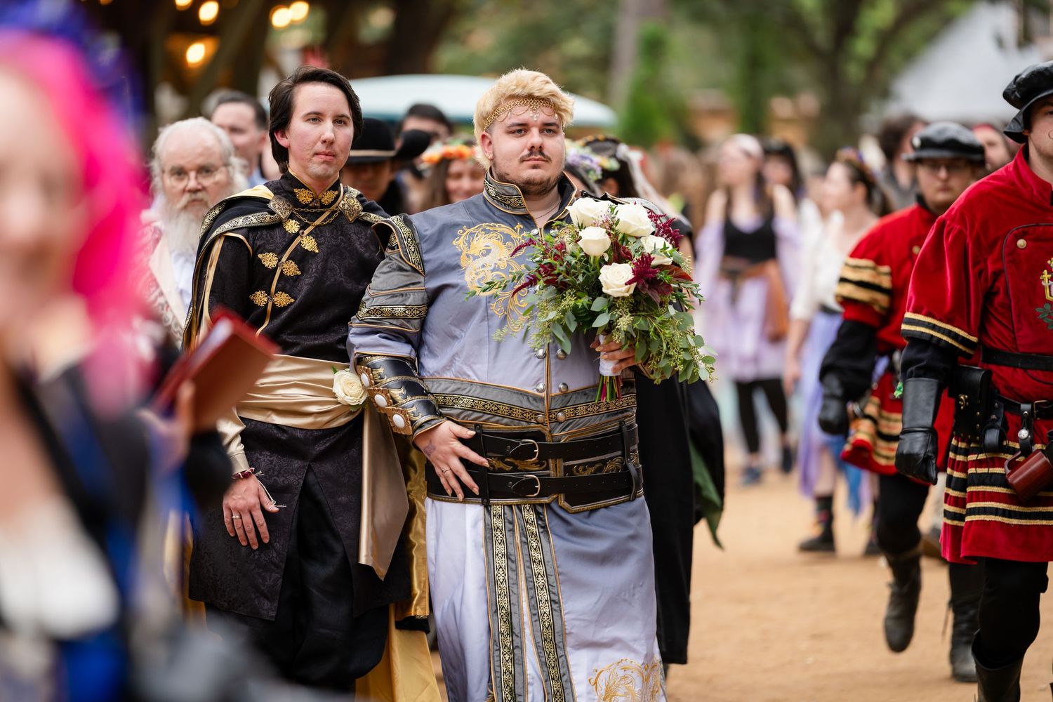 Costumed performers in medieval attire walk through a festival carrying ornate bouquets and wearing detailed period clothing.