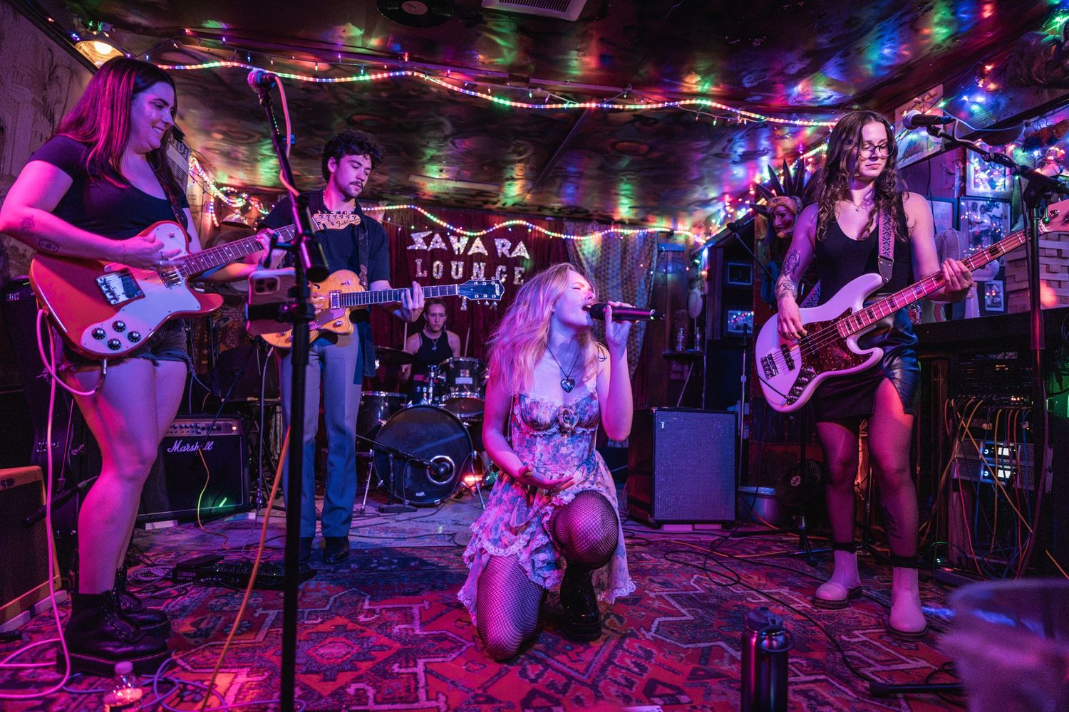 A rock band performs on stage with colorful lighting at a small venue, featuring multiple musicians with instruments.
