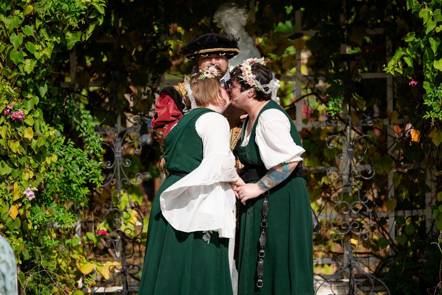 Two people in traditional green and white dirndl dresses embrace in a garden setting with autumn foliage in the background.