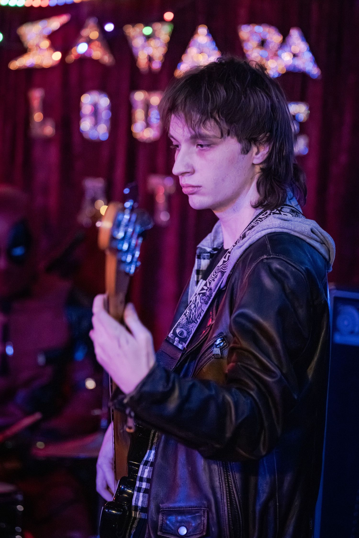 A musician in a leather jacket performs on stage with moody purple lighting in the background.