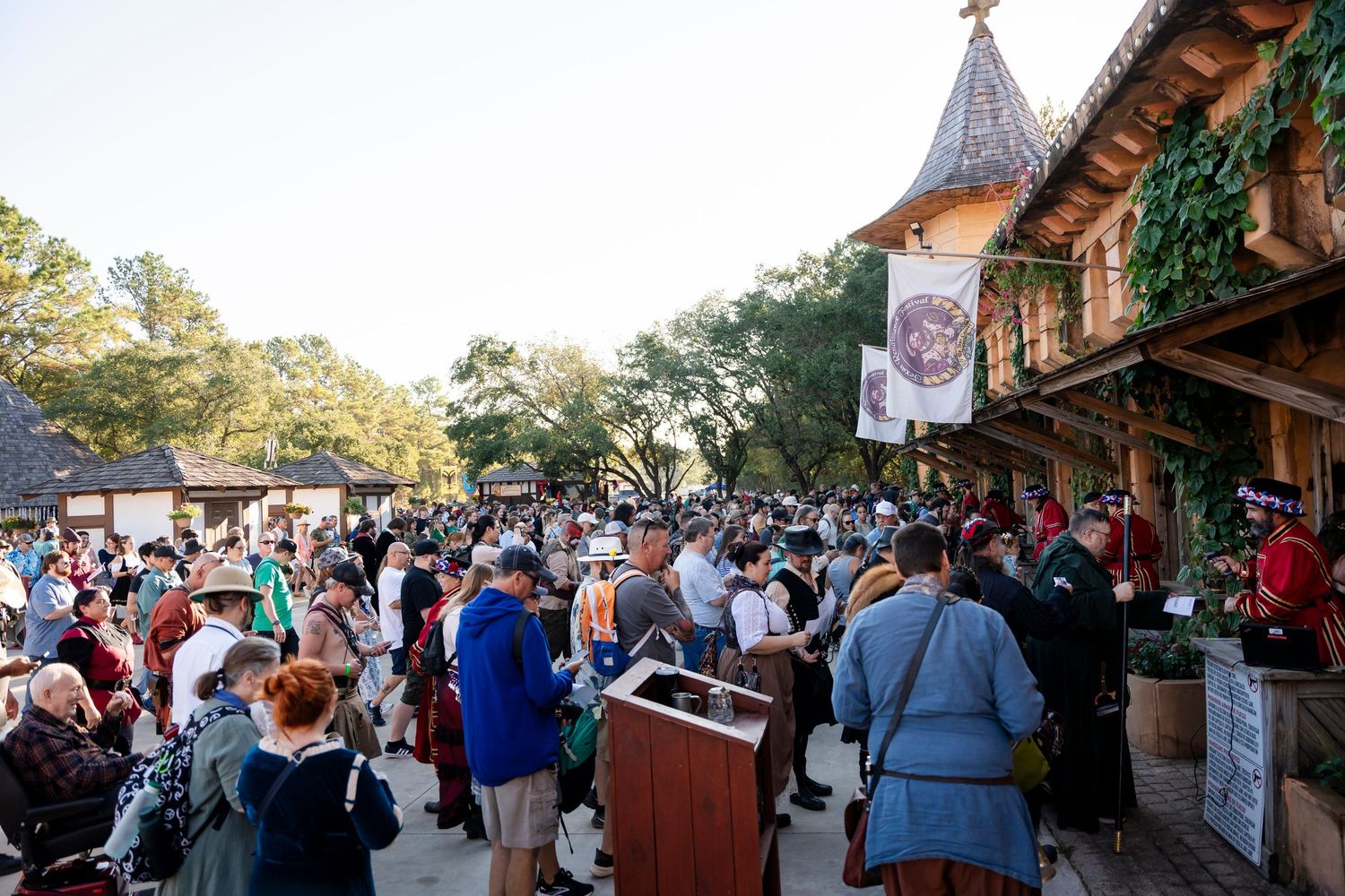 A large crowd gathers at a rustic outdoor market or festival with wooden vendor stalls and autumn decorations.
