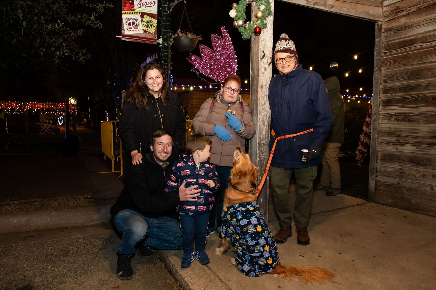 A group stands together for a holiday photo at night with festive lights and decorations in the background.