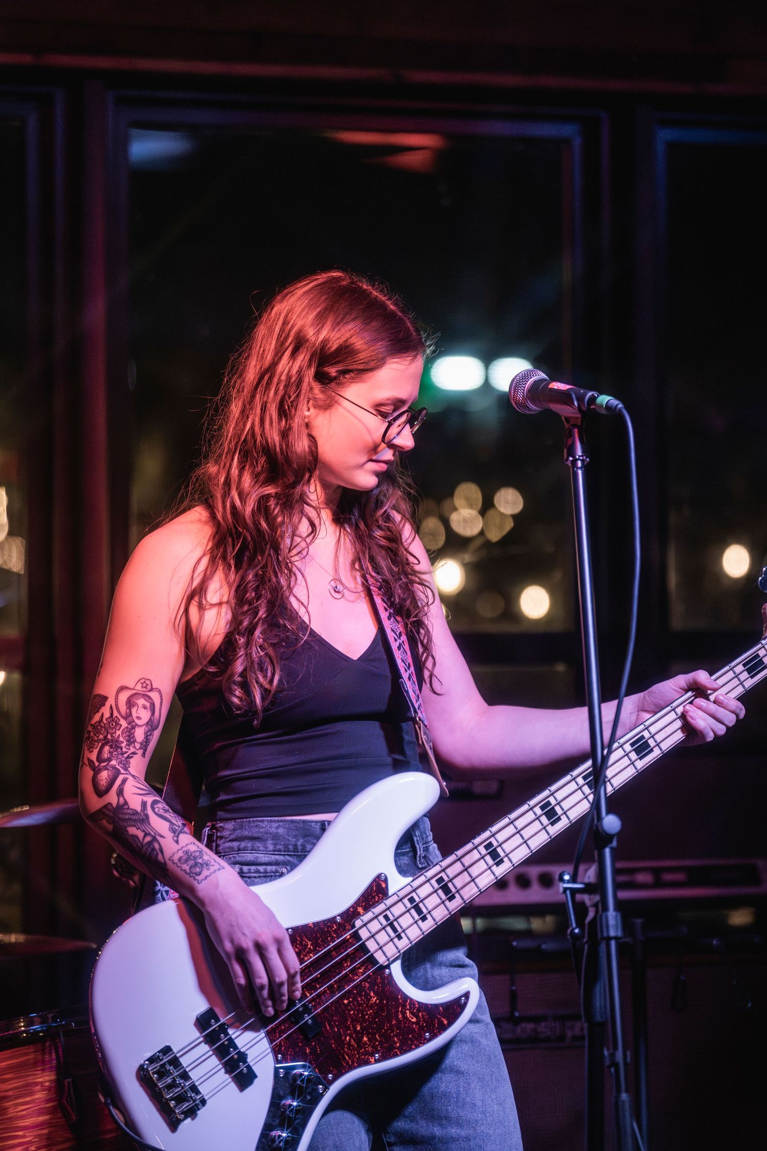 Bass guitarist with tattoos performs on stage in a dark venue with moody lighting and microphone setup.