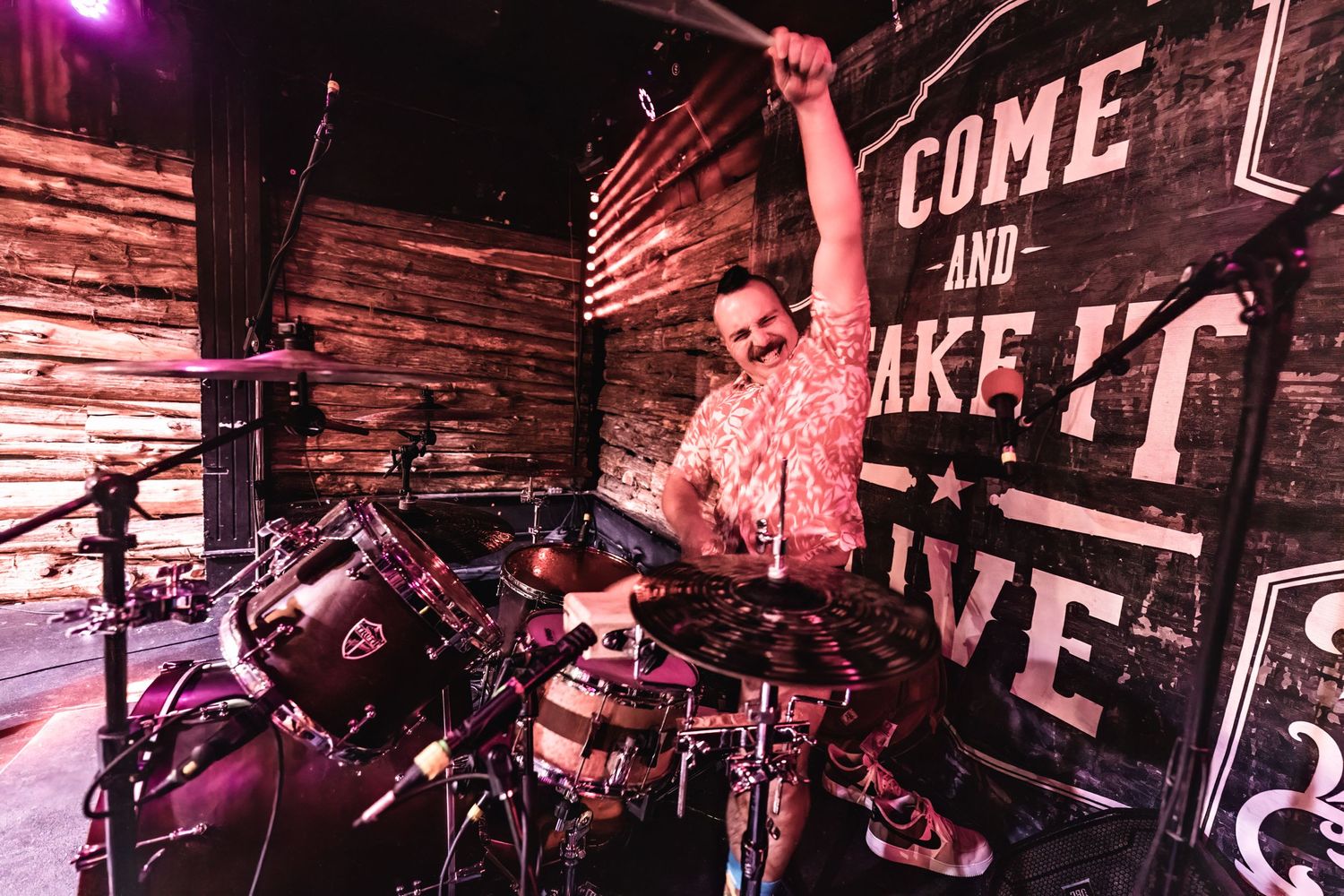 Drummer performs energetically on stage with 'Come and Take It' sign visible in background.