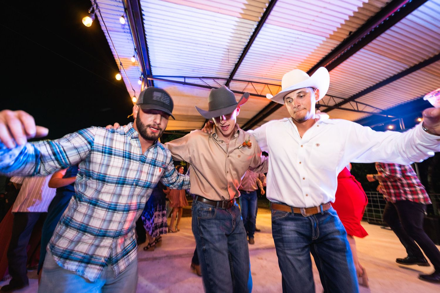 Three men in western wear dancing together at an outdoor party under string lights.
