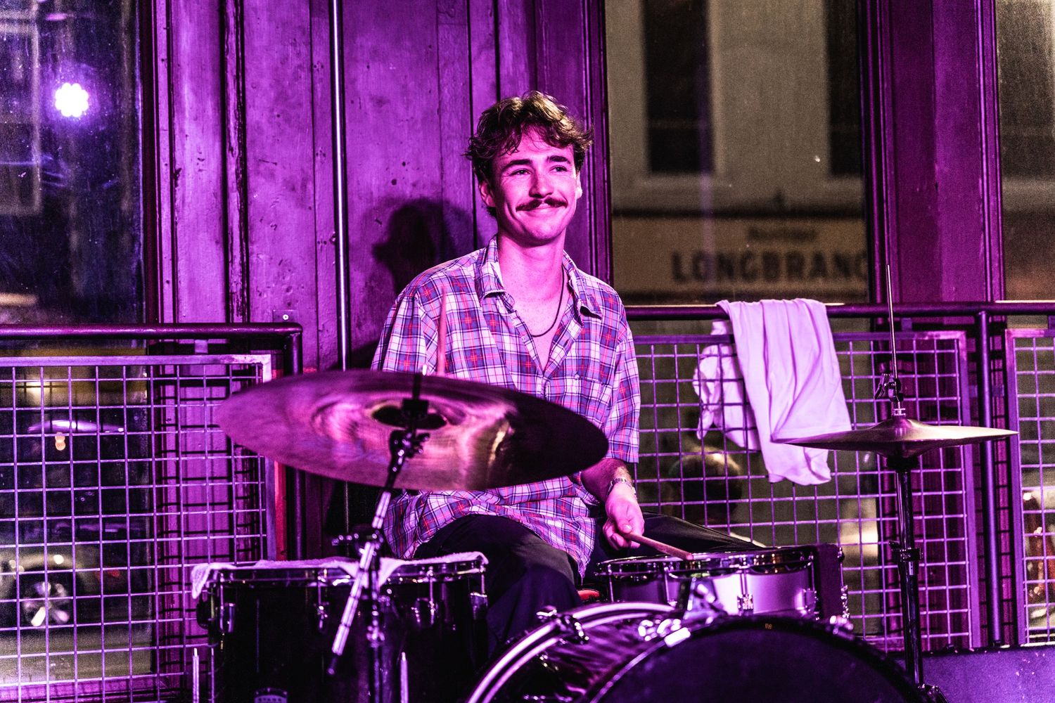 Drummer plays kit on stage illuminated by vibrant purple stage lights.