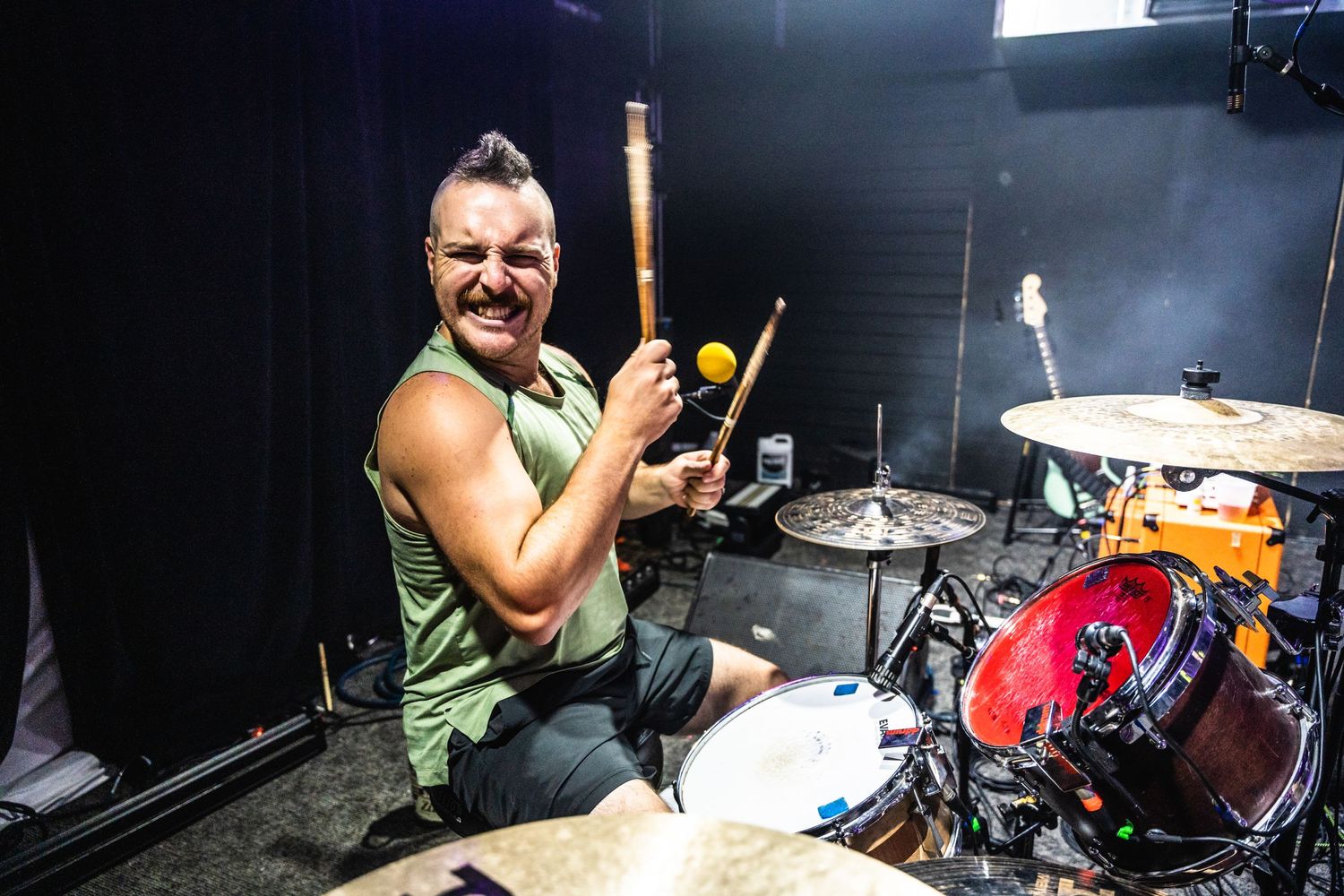 Energetic drummer performs with drumsticks raised while seated at drum kit on stage.