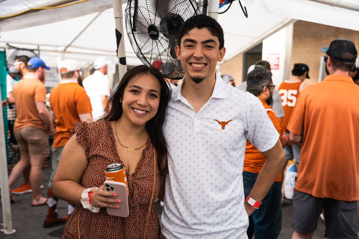Two people smile while holding drinks at outdoor festival with orange-clad crowd in background.