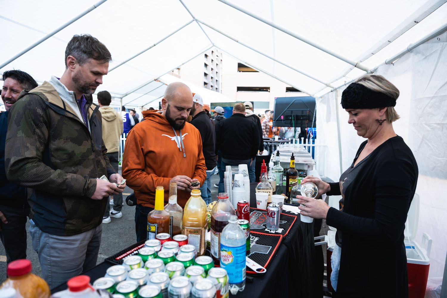 People ordering drinks at an outdoor bar tent with various beverages displayed.
