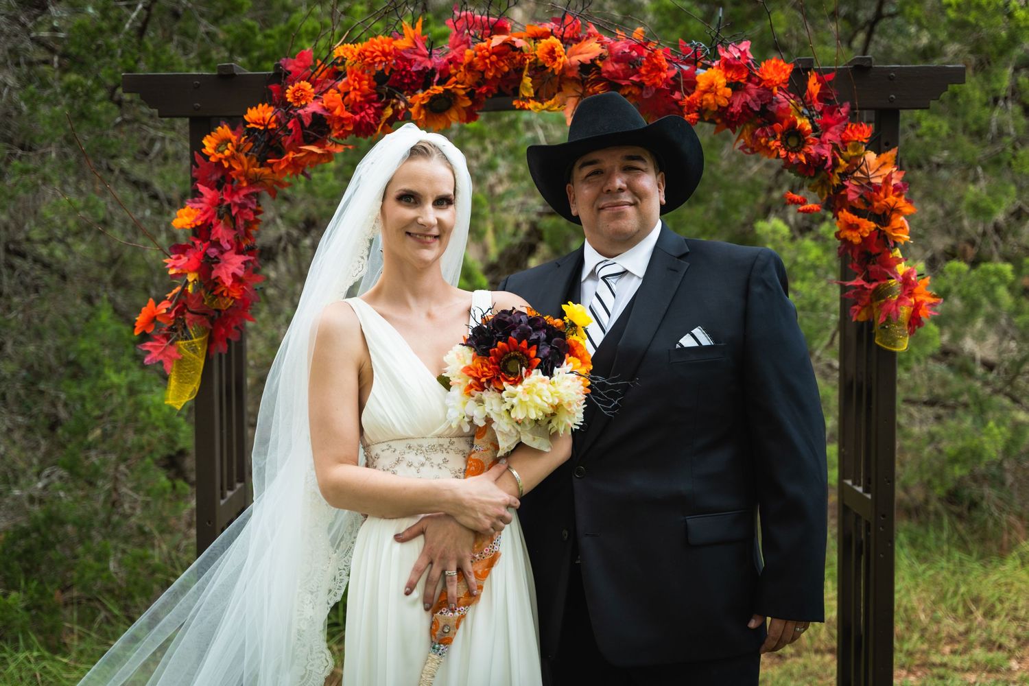 Couple poses under an autumn-colored floral arch with bride holding a fall bouquet.