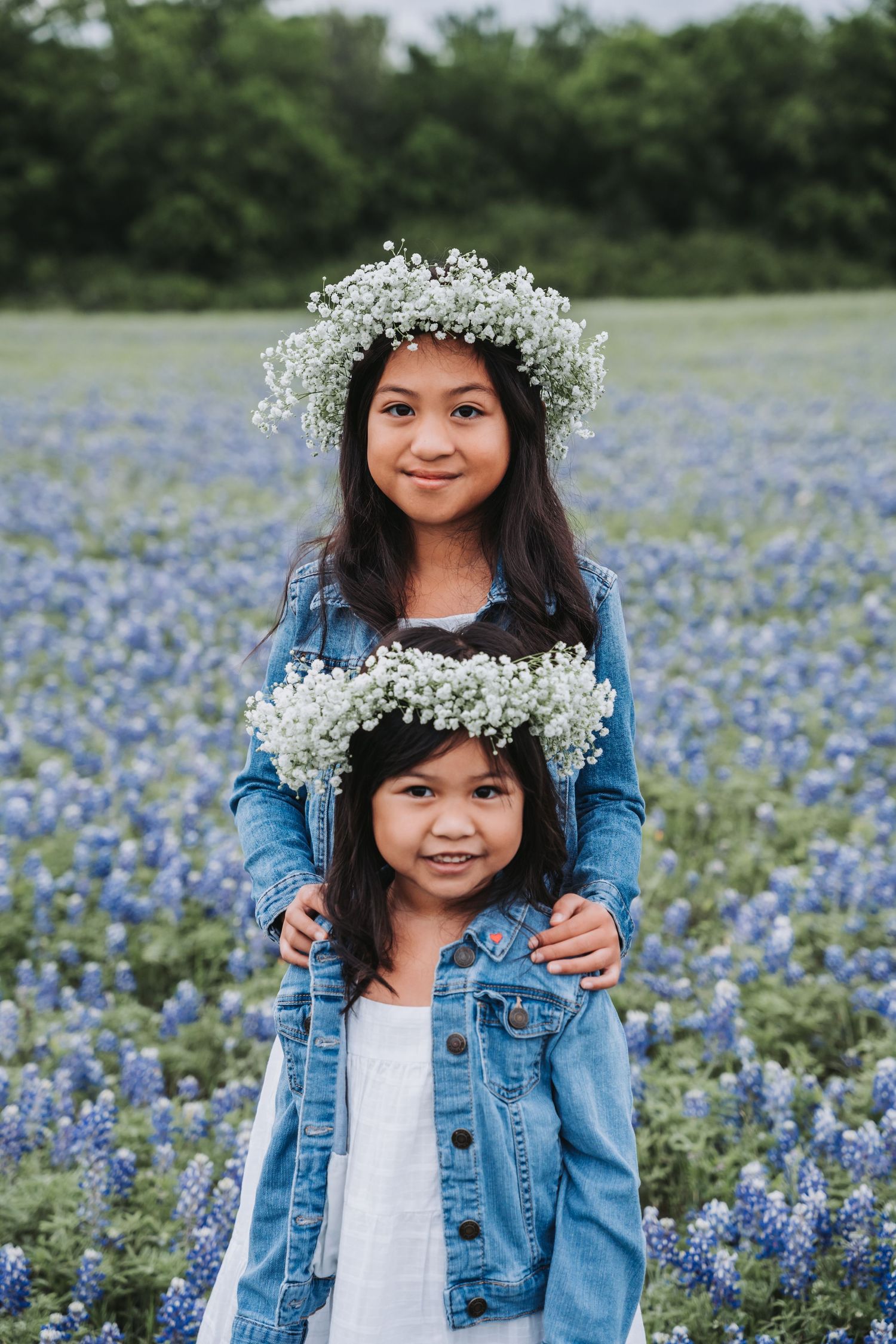 Two figures wearing flower crowns stand together in a field of blooming bluebonnets.