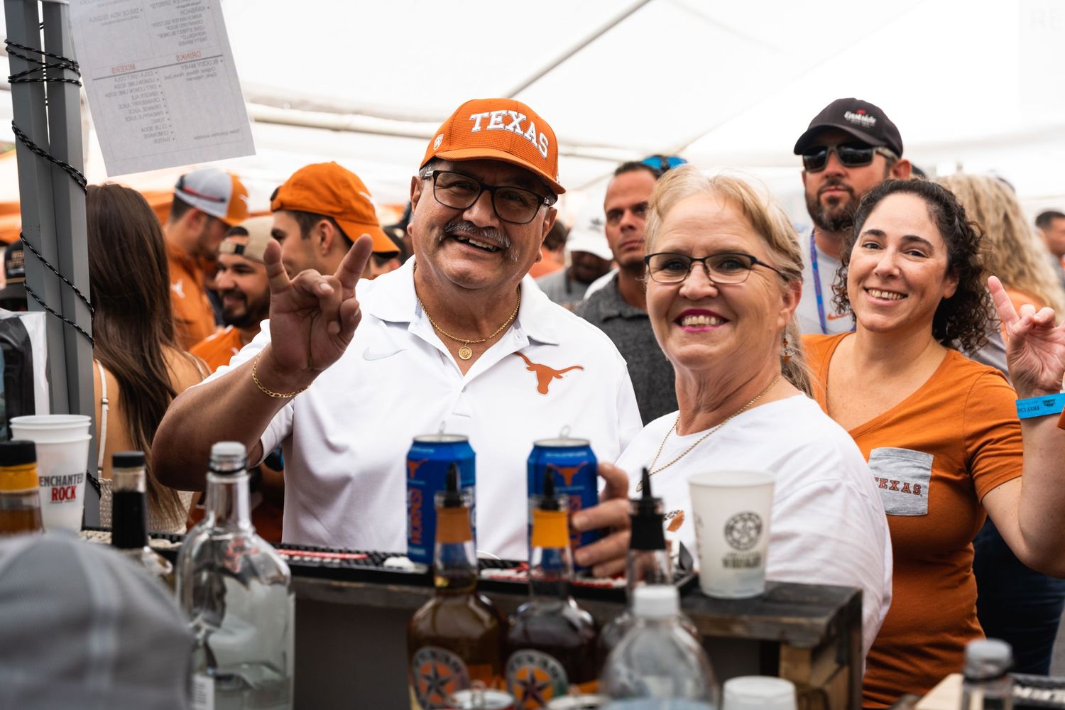 People serving drinks and socializing at outdoor bar under white tent.