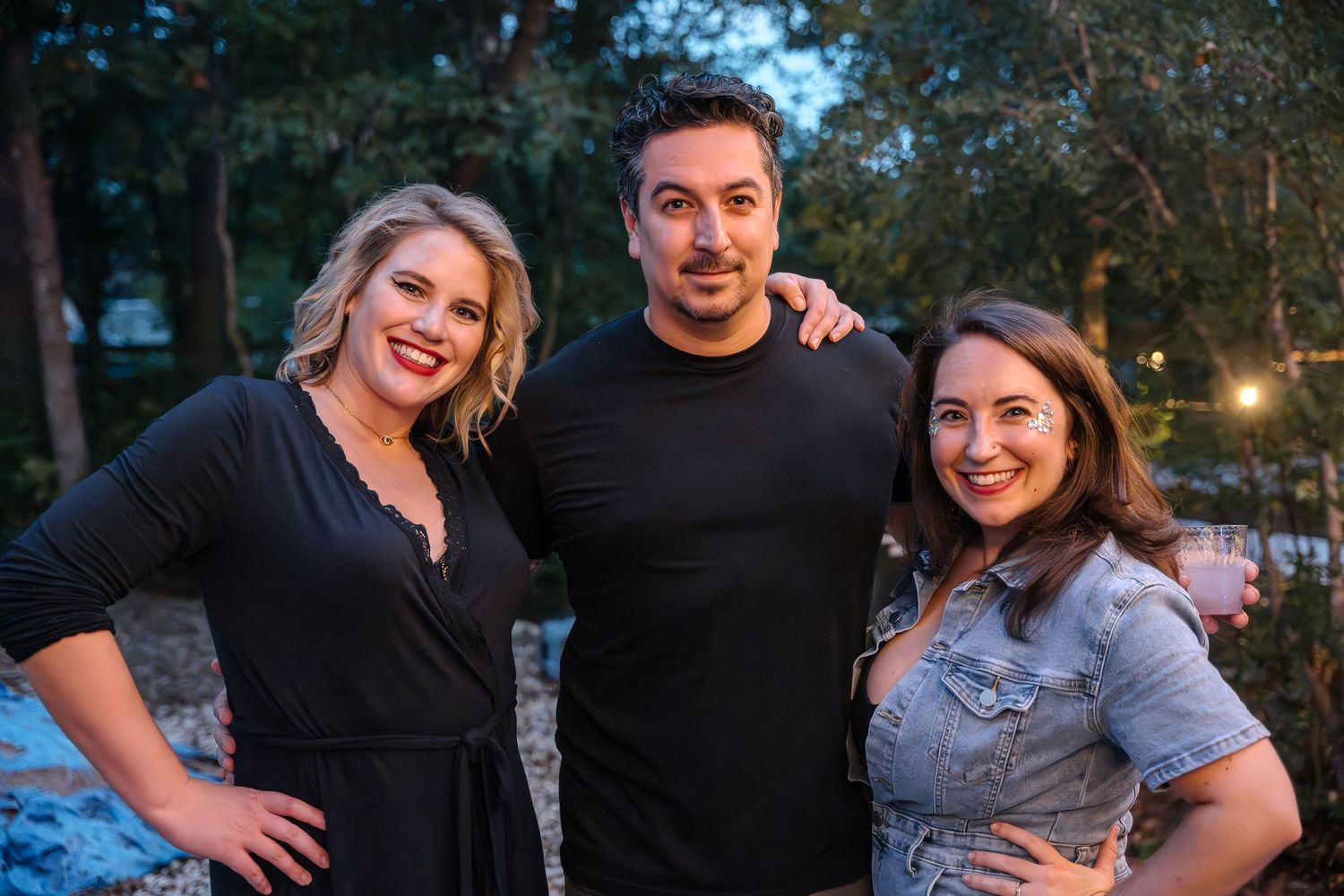 Three people posing together at an outdoor evening party with string lights.