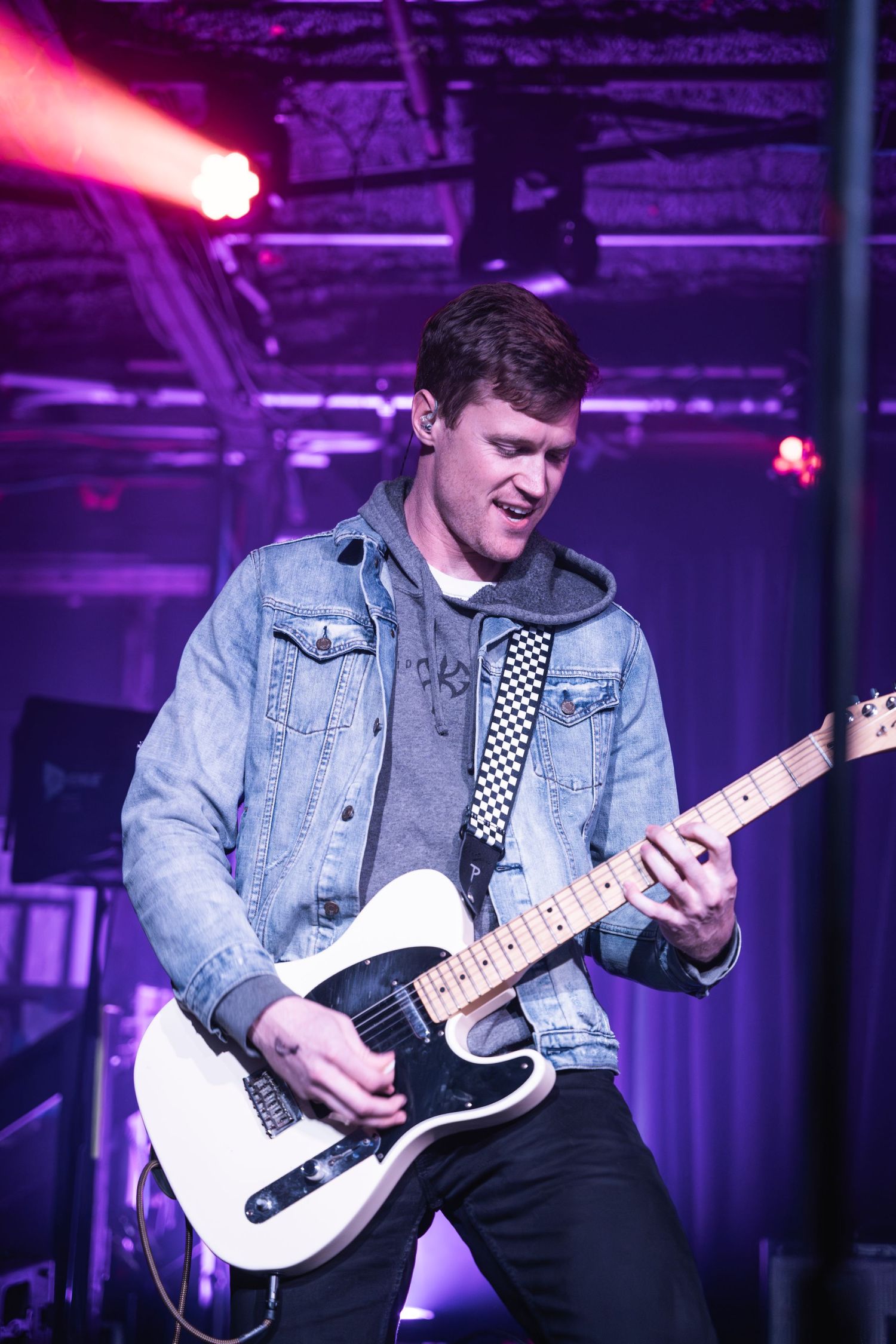 Guitarist playing white telecaster under purple stage lighting wearing denim jacket.
