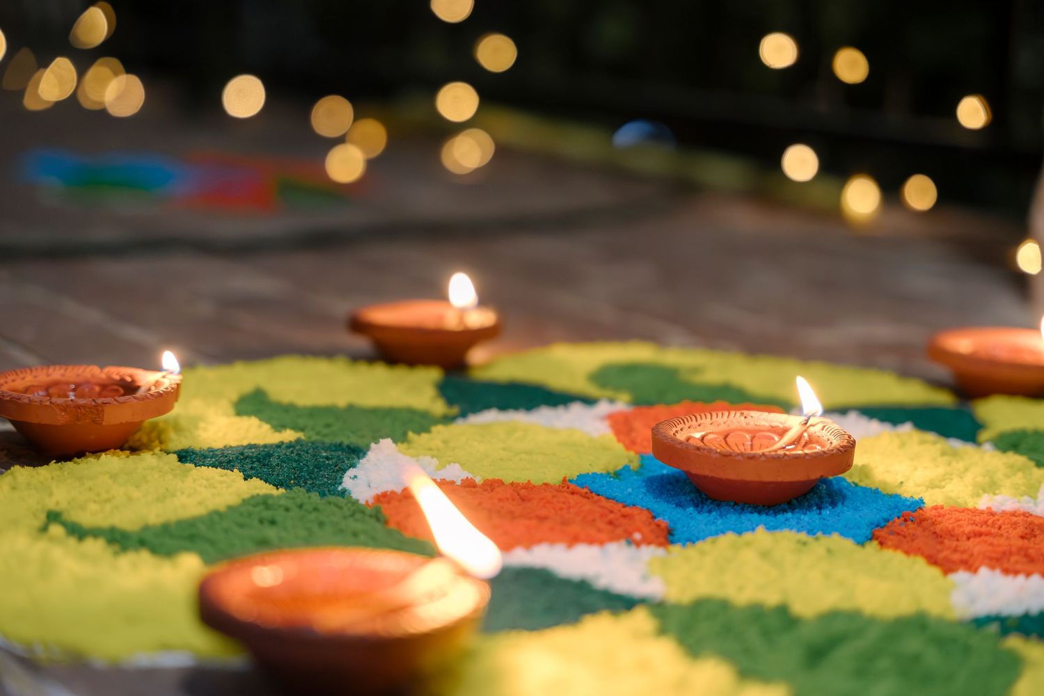 Lit diyas placed on a colorful rangoli design with bokeh lights in the background.