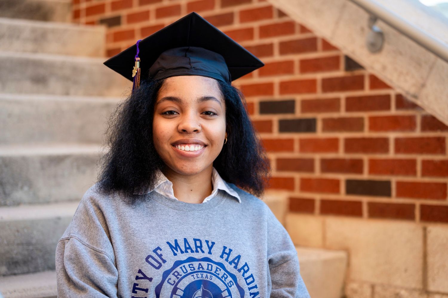 A graduate wearing a University of Mary Hardin-Baylor sweatshirt poses with cap and gown.