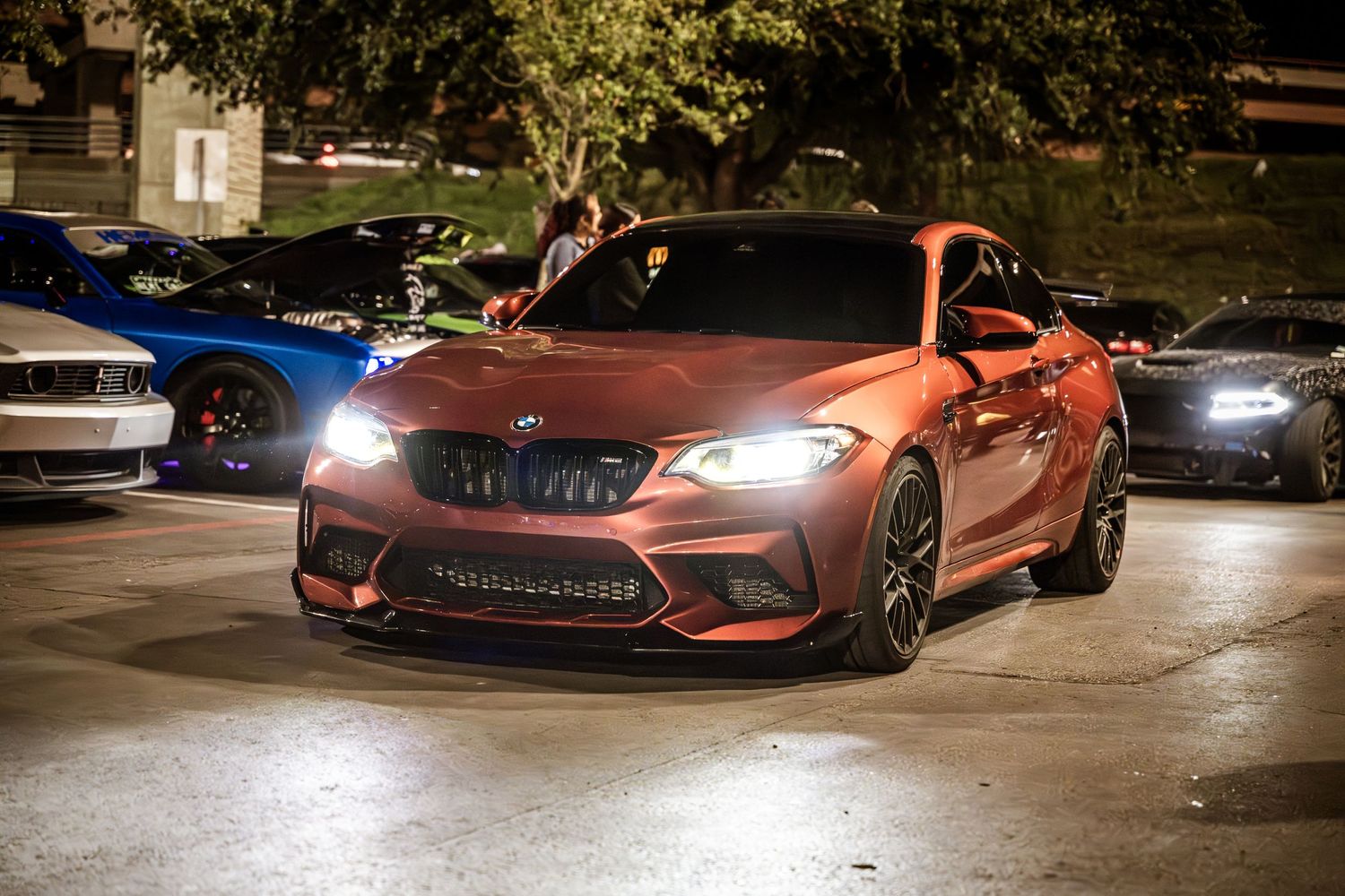 Red BMW M2 sports car with glowing headlights at night car meet.