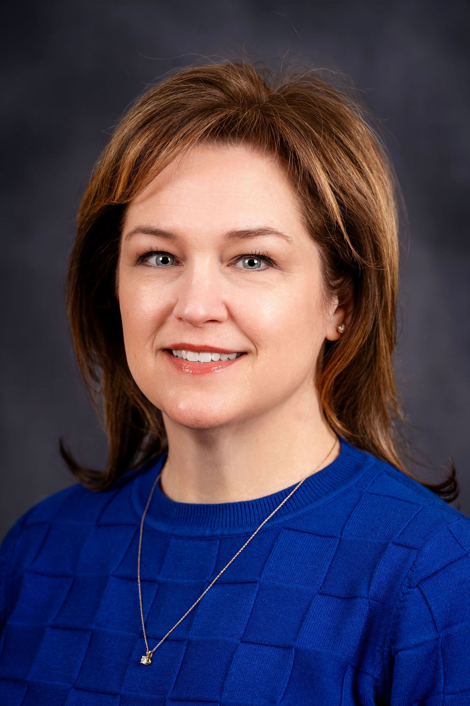 Professional portrait of a person in a blue textured sweater with delicate necklace against dark backdrop.