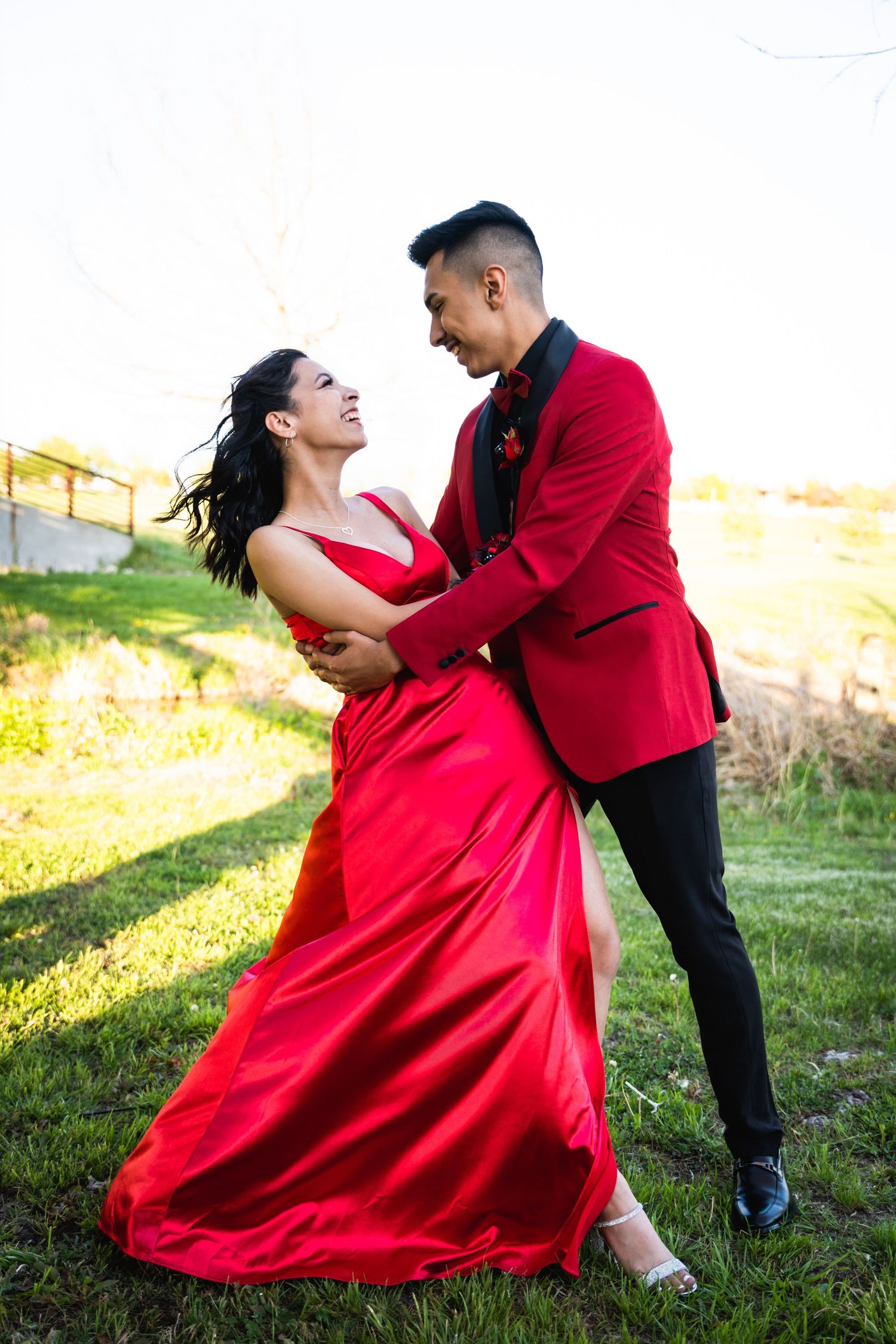 A romantic couple in red formal wear dance together on a grassy hill at sunset.