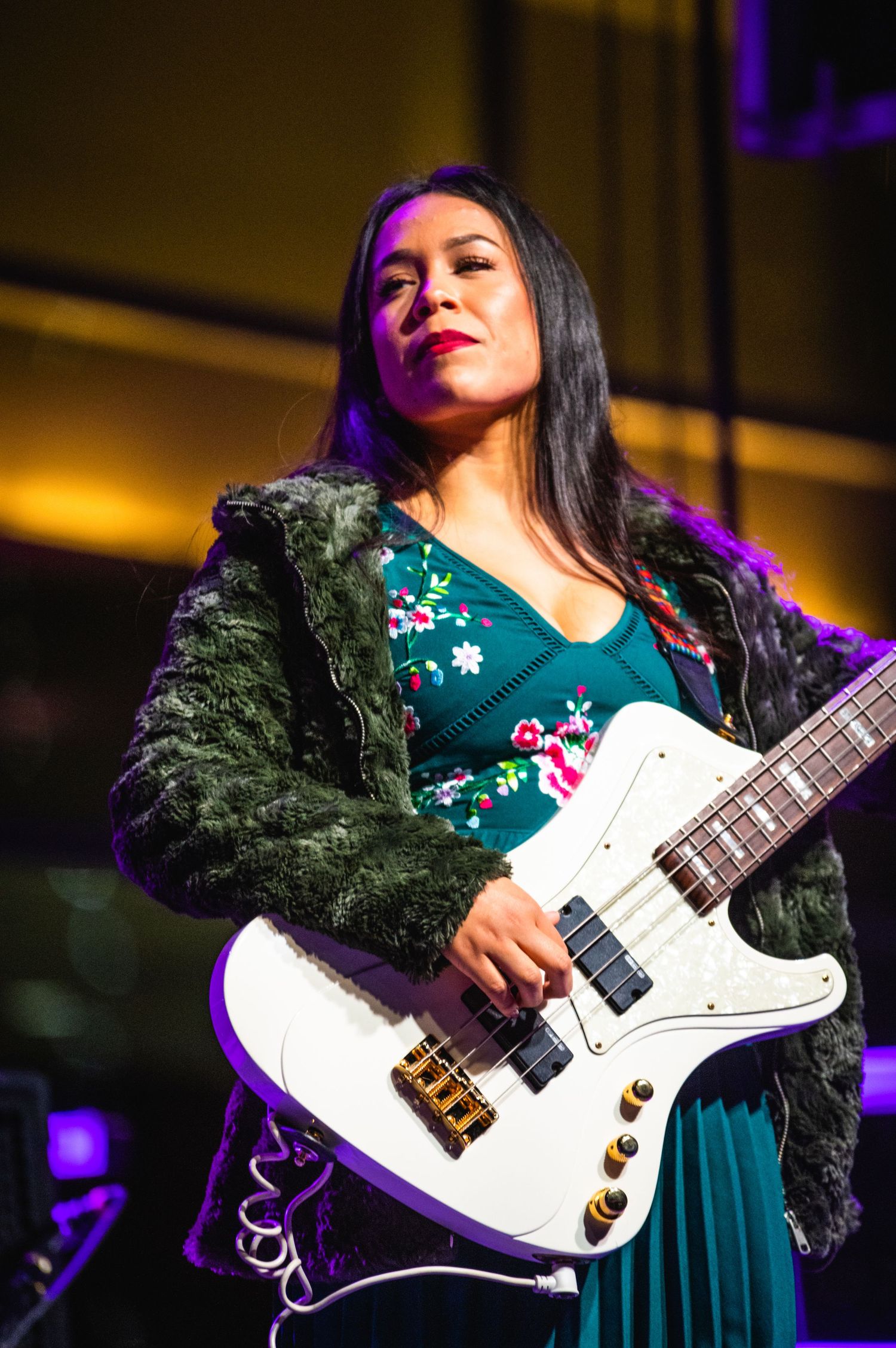 A guitarist performs on stage wearing a green floral dress and fuzzy black jacket while playing a white electric guitar.