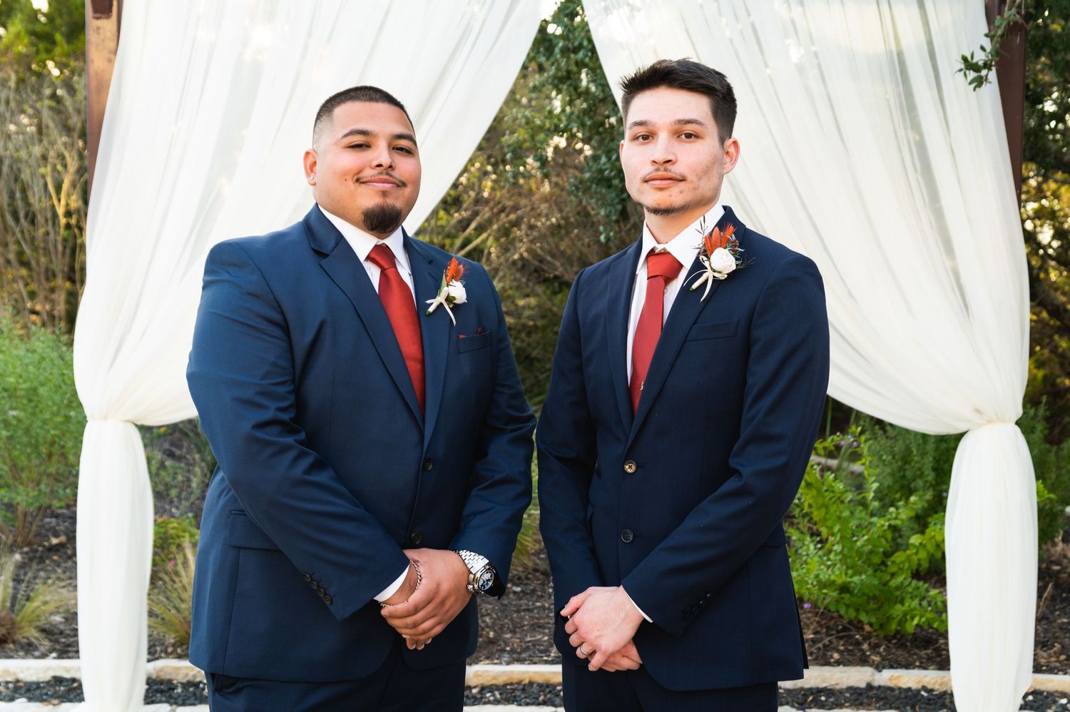 Two groomsmen in navy suits with red ties stand together under white wedding ceremony drapes.