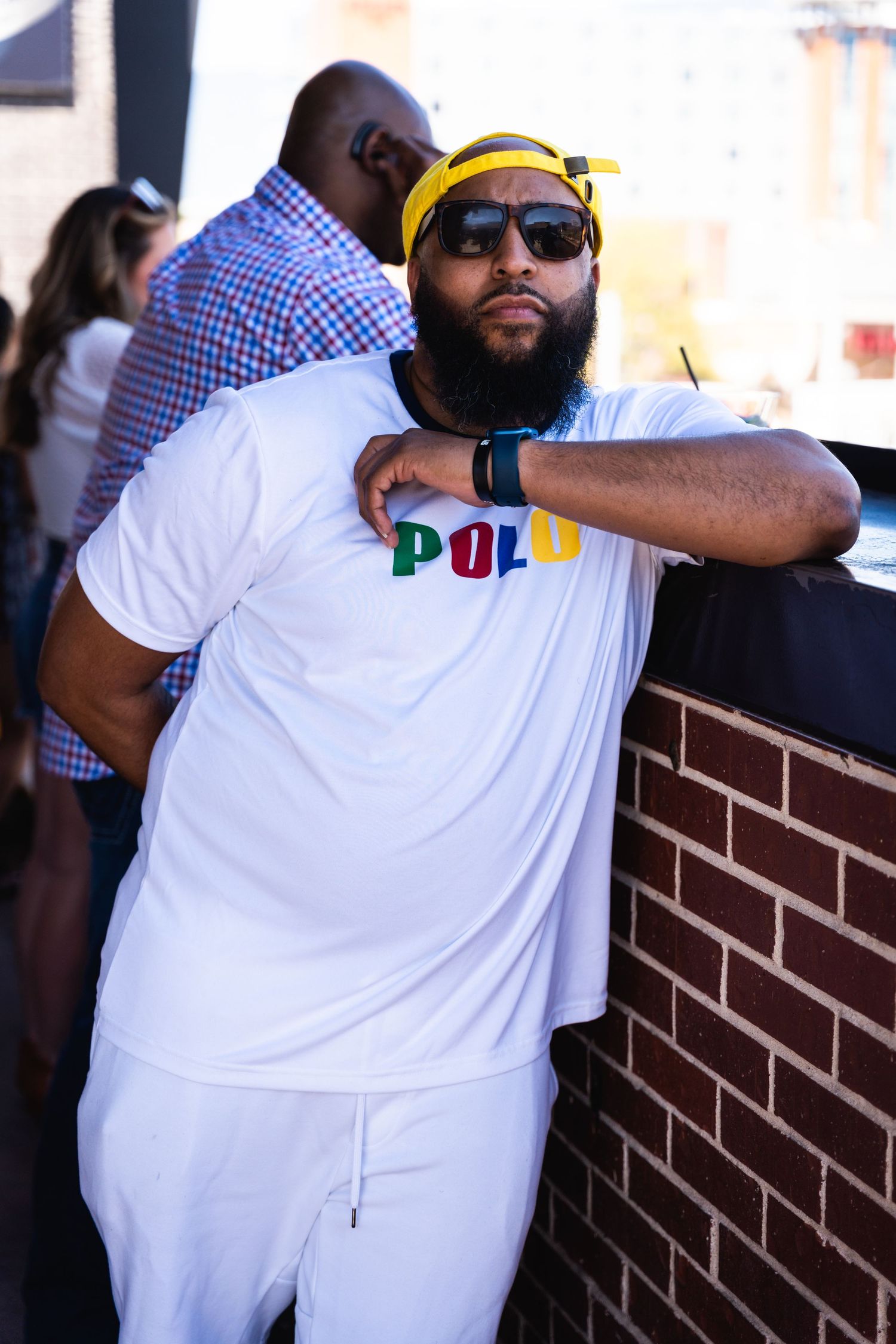 Person in white Polo t-shirt and yellow cap leaning against brick wall in casual pose.