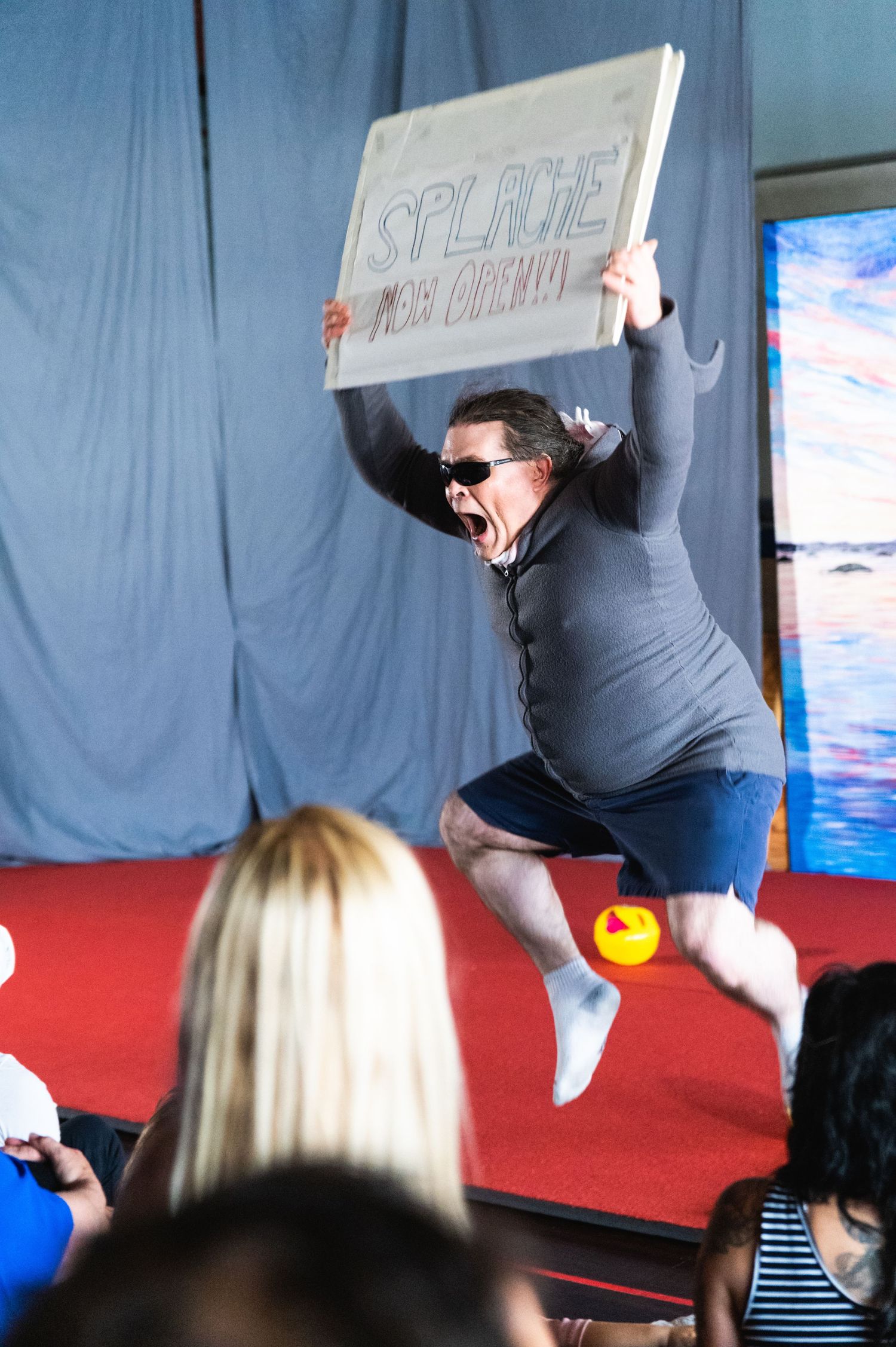 Performer jumps energetically while holding sign during indoor performance.