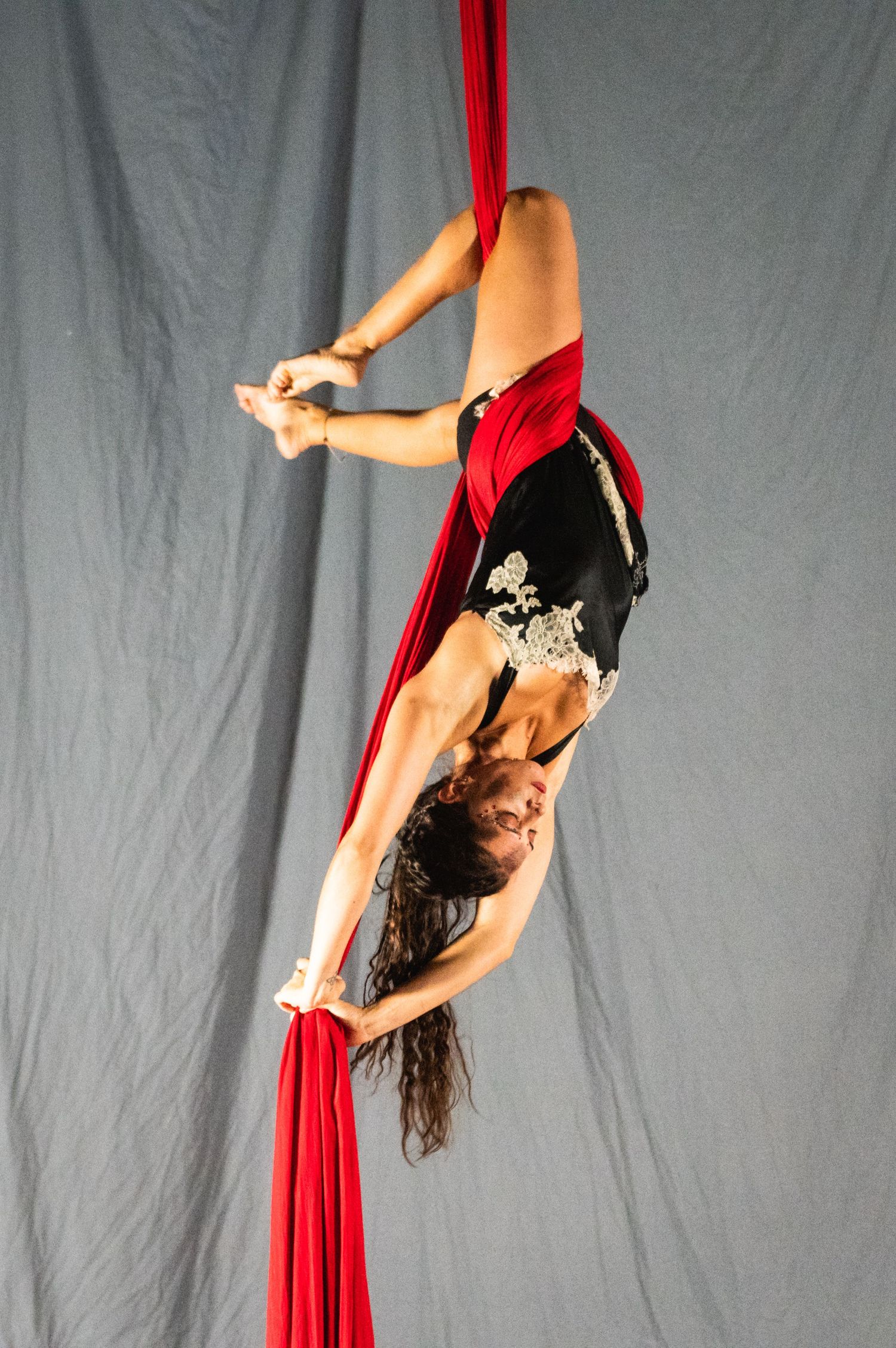 Aerialist in black costume performs an inverted pose while suspended on red silk fabric.