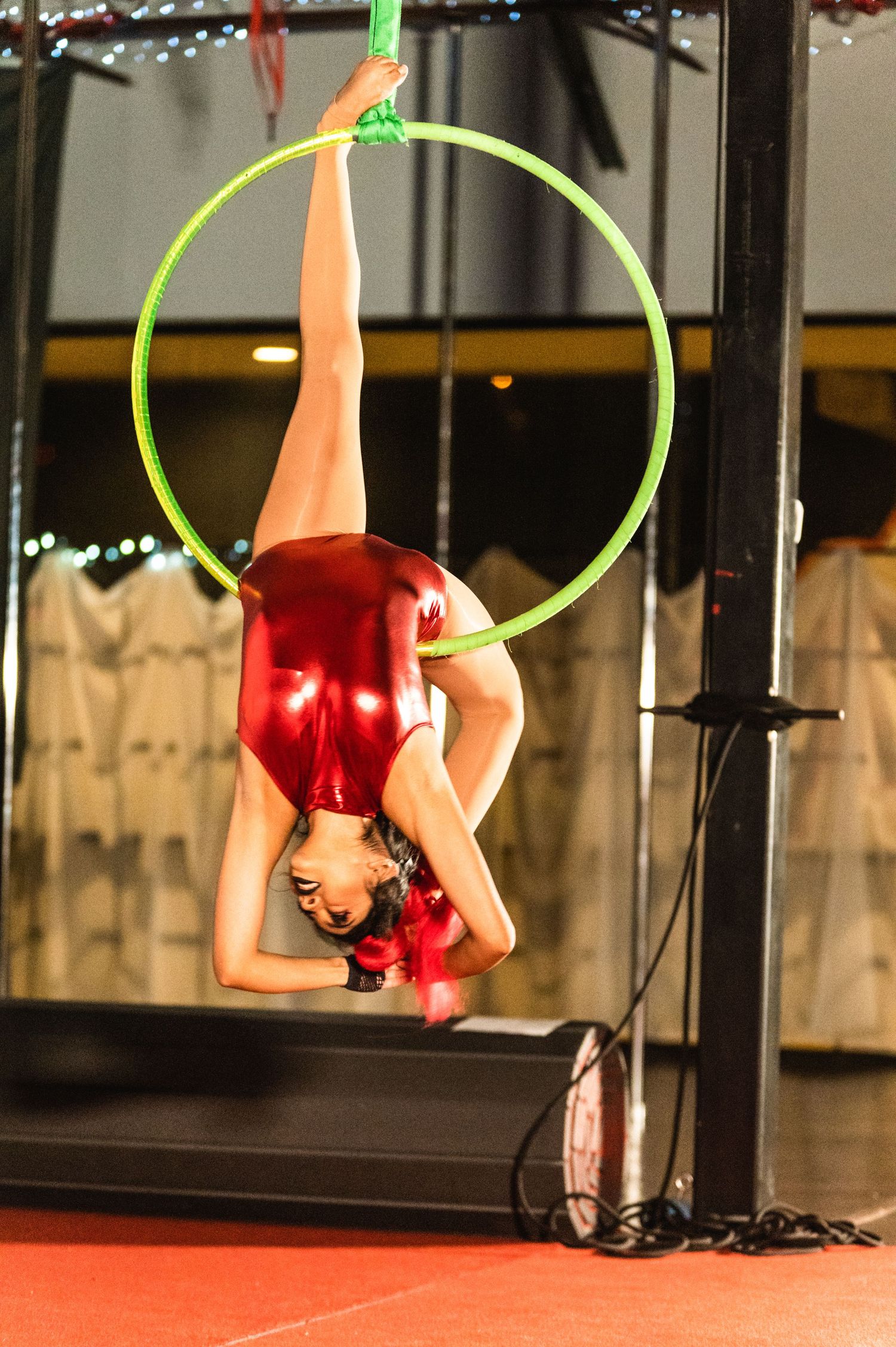 A performer in a red metallic costume demonstrates strength and flexibility on a green aerial hoop.