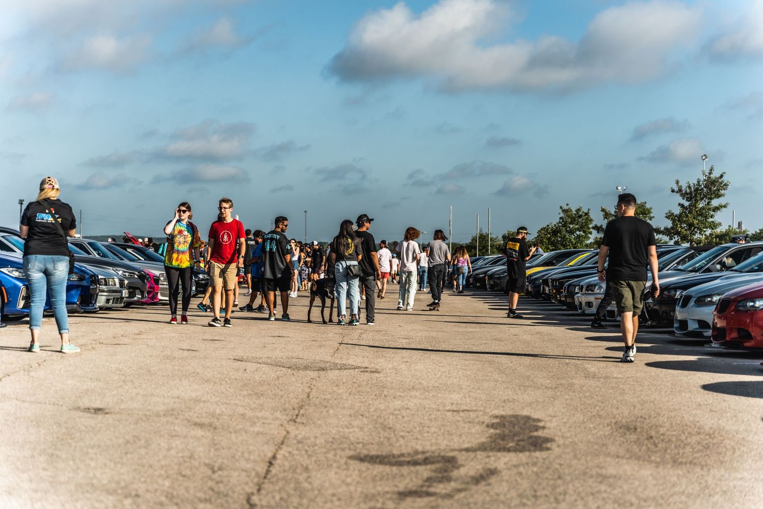Crowd walking through outdoor car show parking lot on sunny day.