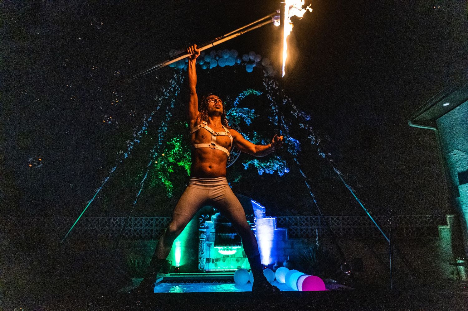 Fire performer executes dramatic pose with flaming props against colorful lighting display.