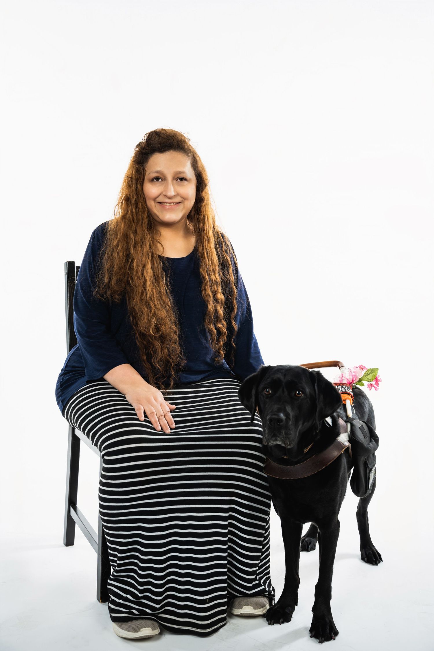 Woman in striped skirt sitting with black Labrador service dog on white background.