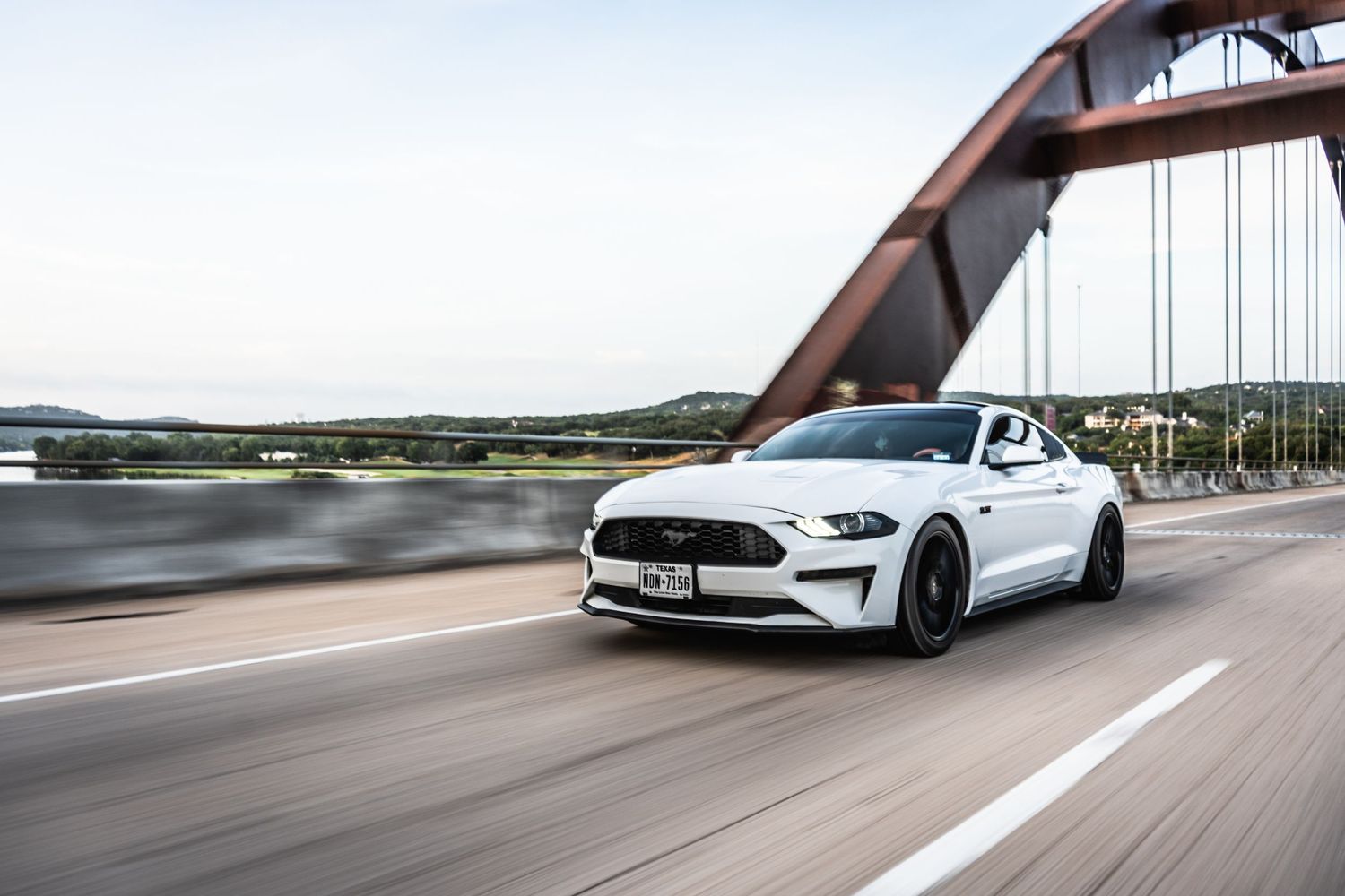 White Ford Mustang sports car driving across a bridge with dramatic architecture.