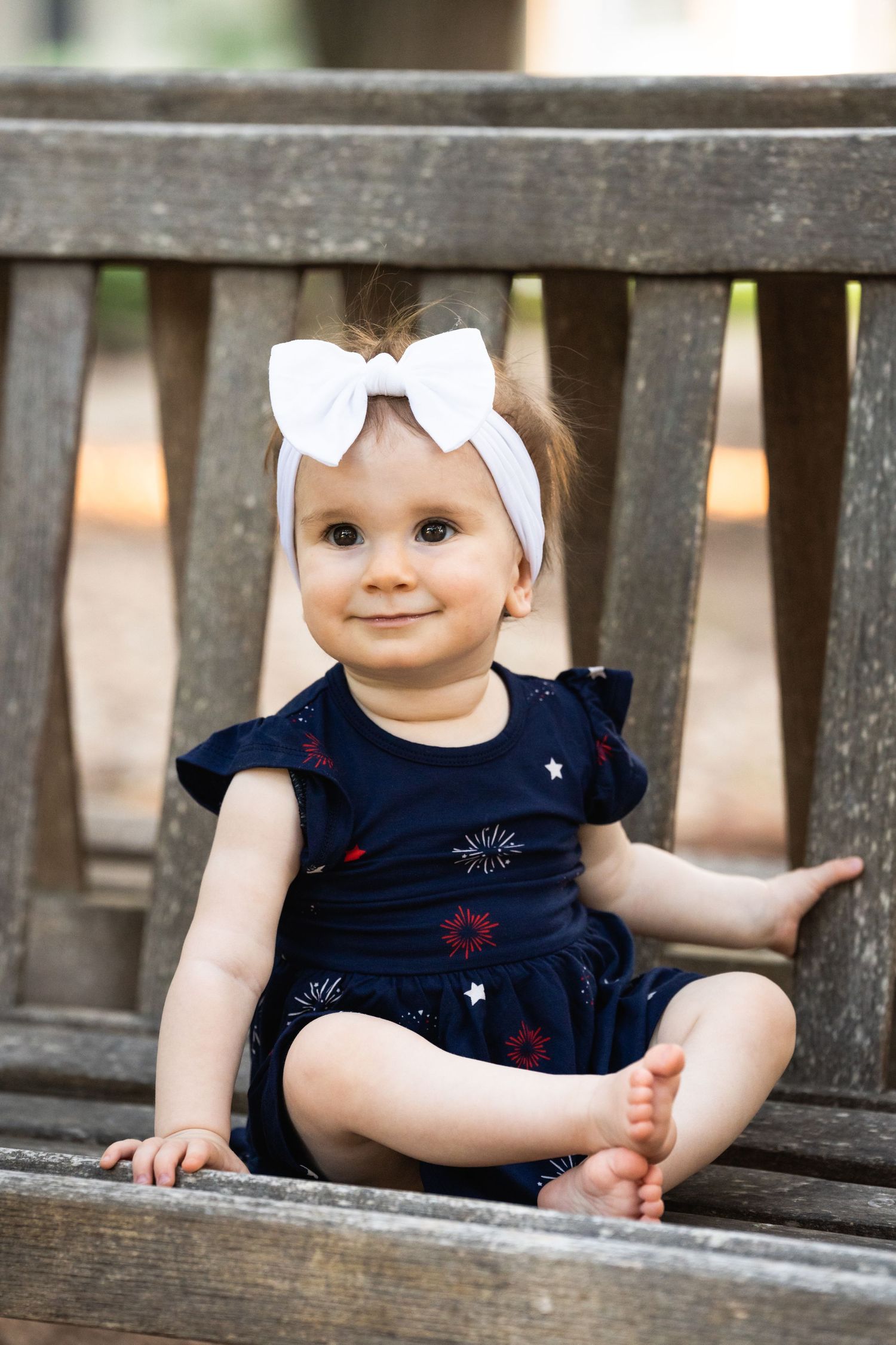 Small child sits on wooden bench wearing navy dress and white bow headband.