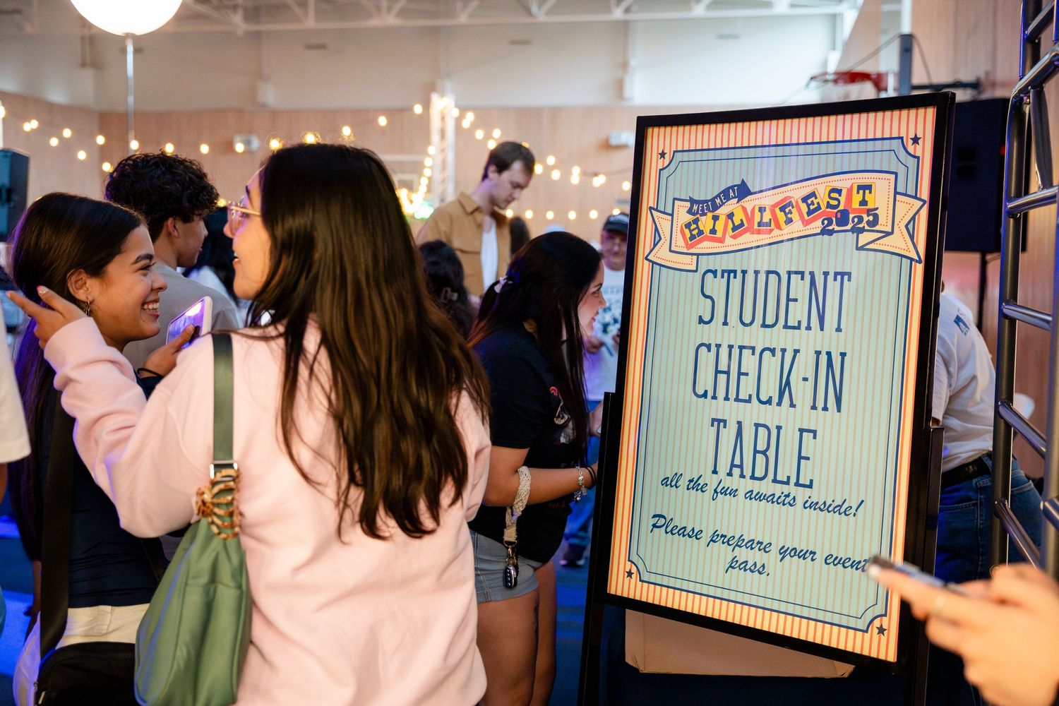 A student check-in table sign at an event with string lights and blurred attendees in the background.
