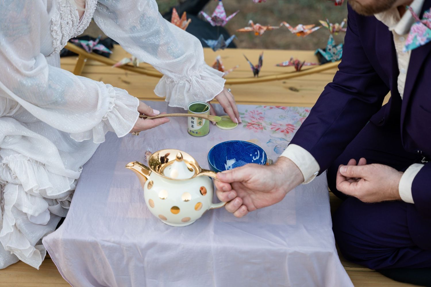 A traditional tea ceremony takes place at an outdoor wedding with an ornate teapot on a white tablecloth.