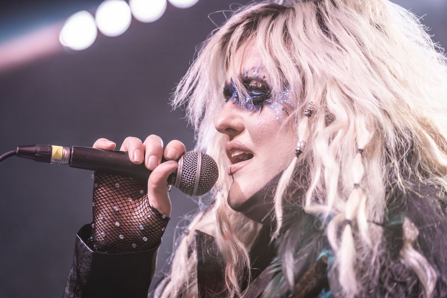 Concert performer with bright blonde hair sings passionately into microphone under stage lights.