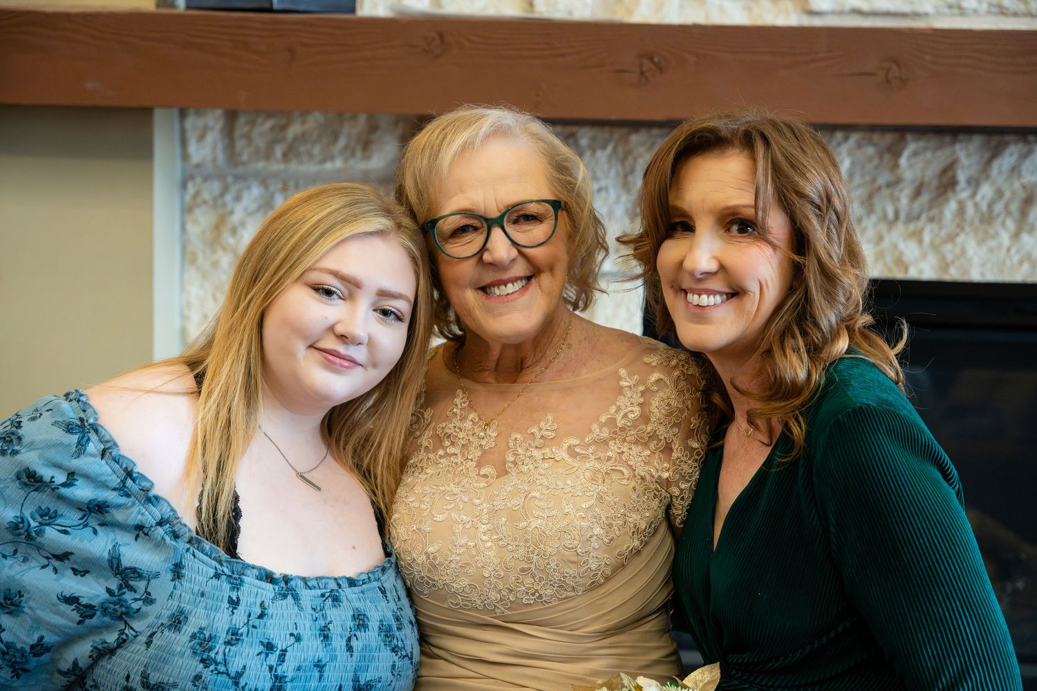 Three generations of women smiling together while wearing formal attire in front of a fireplace.