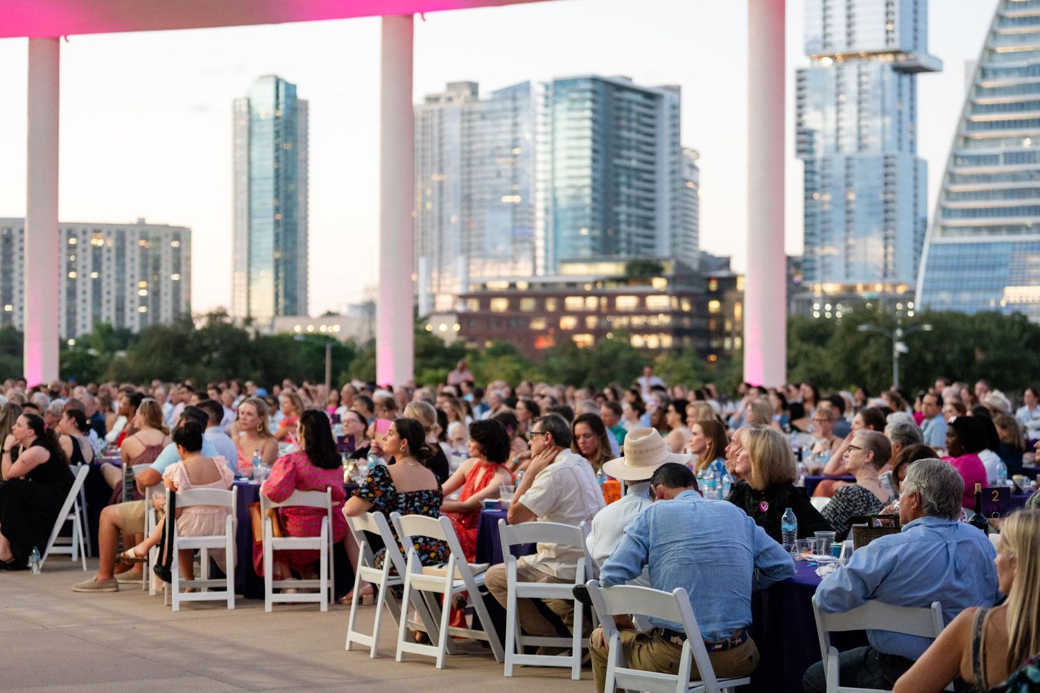 Large crowd seated at outdoor tables during an evening event with city skyline visible in the background.