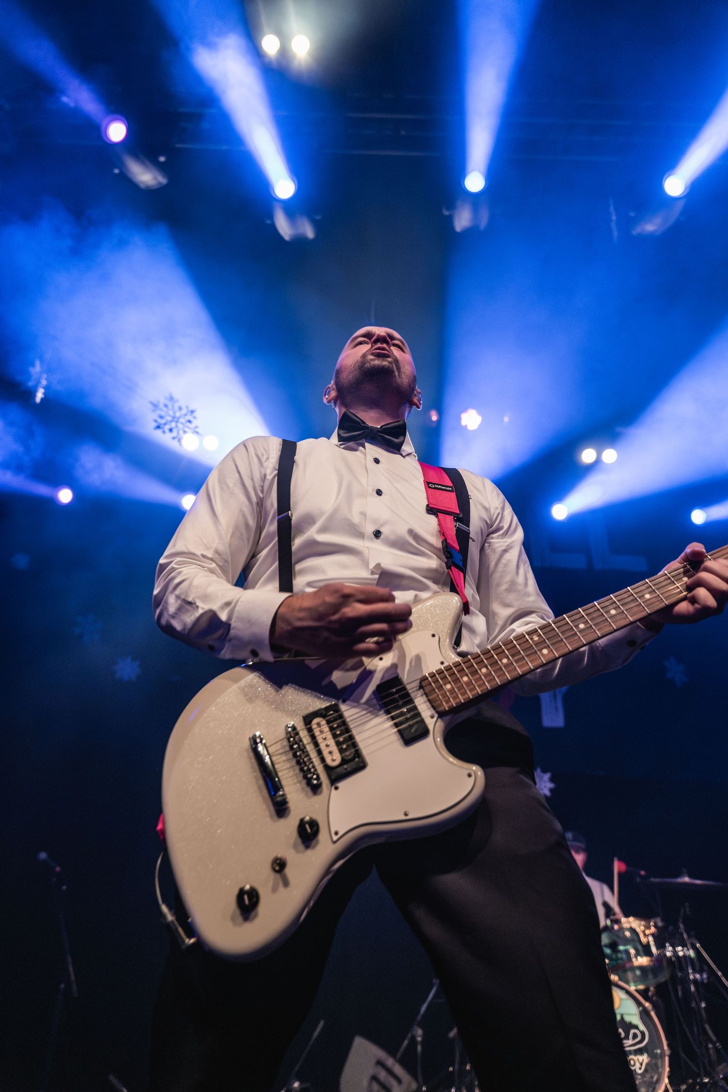 Guitarist in white shirt and suspenders performing passionately under blue stage lights.