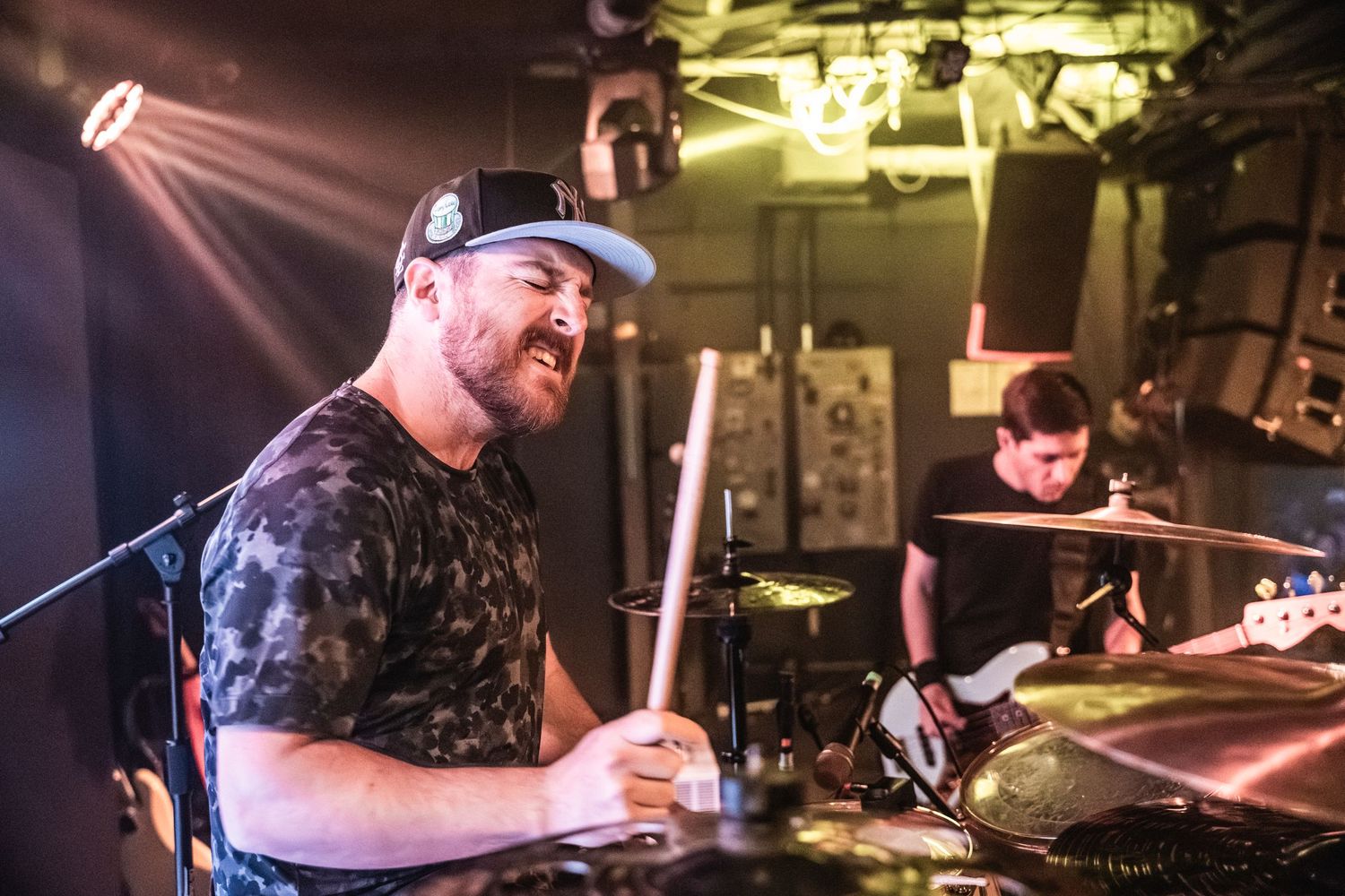 A musician performs with drumsticks on stage during a live performance with colorful lighting effects.