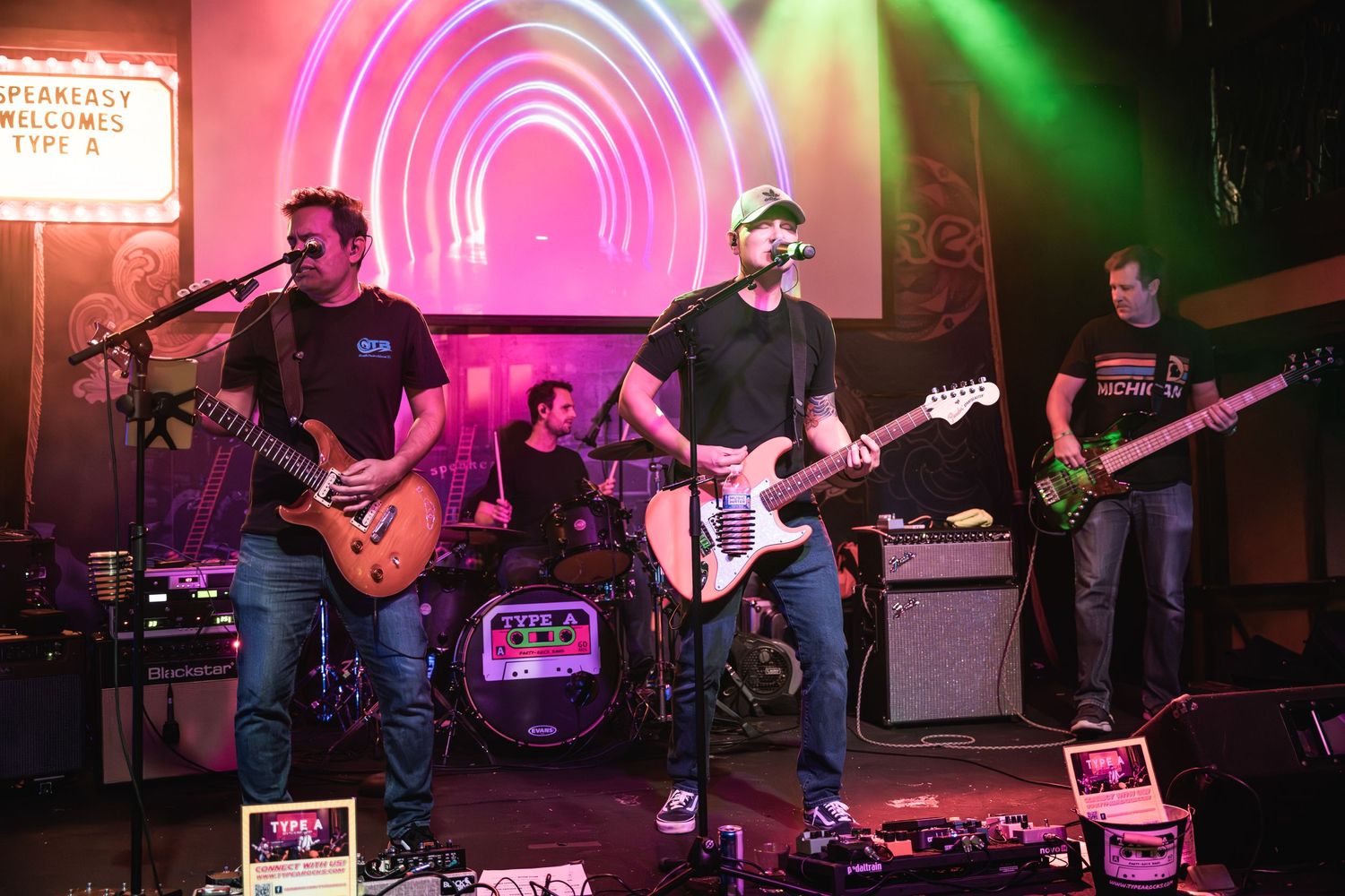 Rock band performs on stage with colorful pink and green lighting effects backdrop.