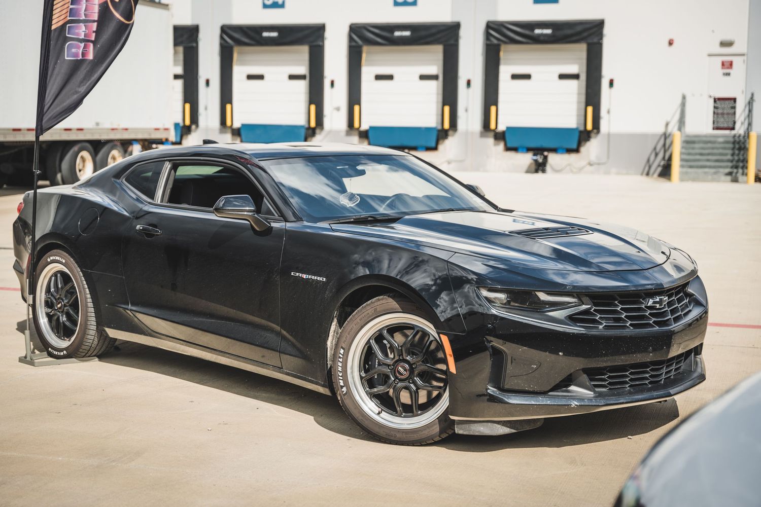 Black Chevrolet Camaro with custom wheels positioned near warehouse loading bays.