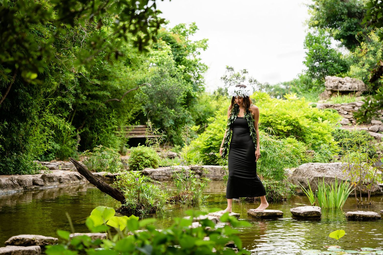 Figure in black dress stands on stones near tranquil pond in lush garden setting.