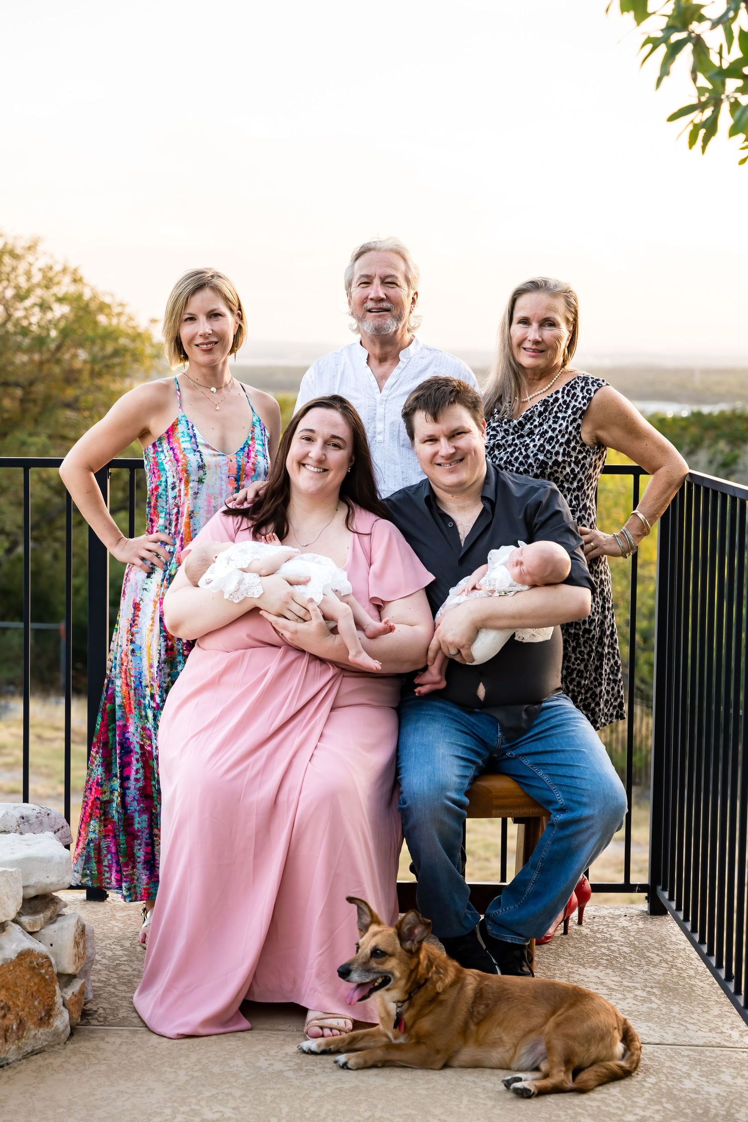 Family portrait on outdoor deck with twins, dog, and scenic background at sunset.