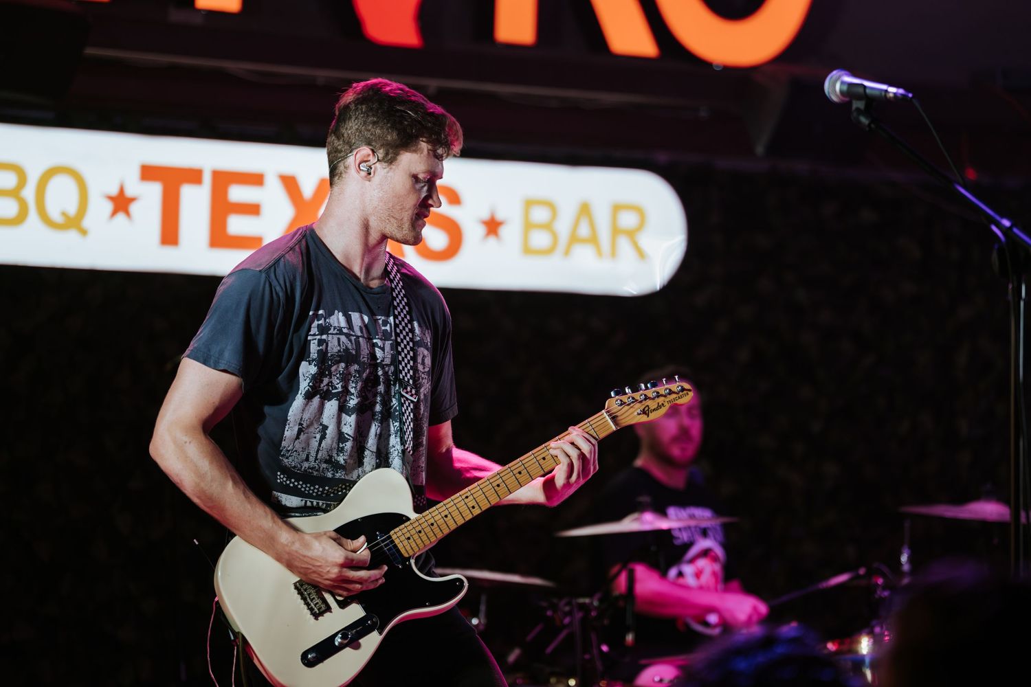 Guitarist playing white electric guitar on stage with BBQ Texas Bar sign in background.