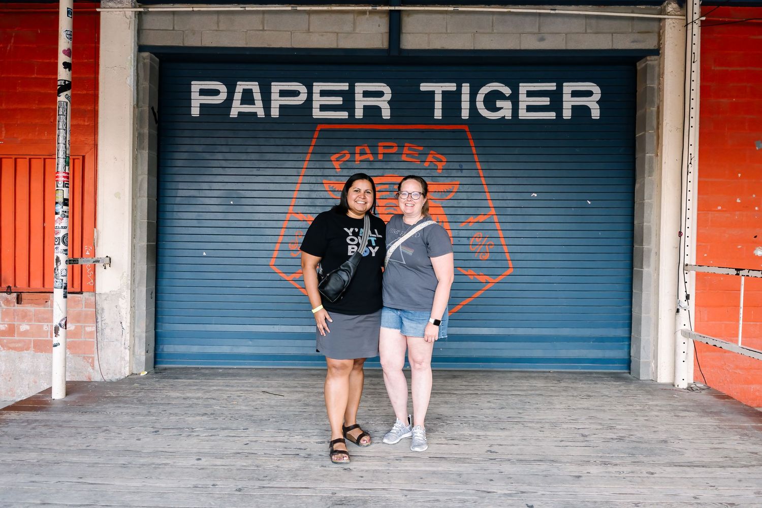 Two people pose in front of a closed Paper Tiger storefront with blue metal gate.