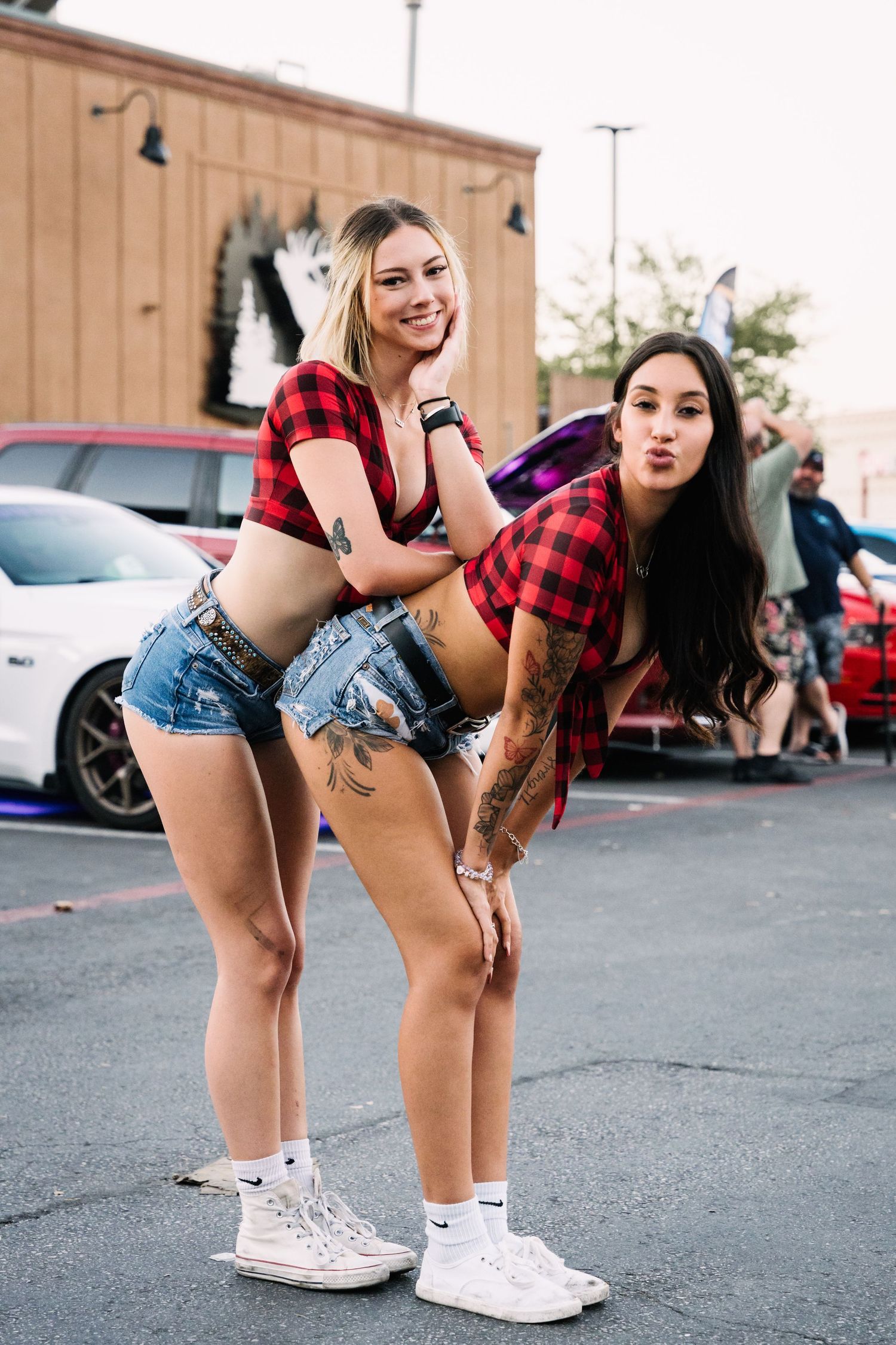 Two models in matching red plaid shirts and denim shorts pose playfully at a car meet.