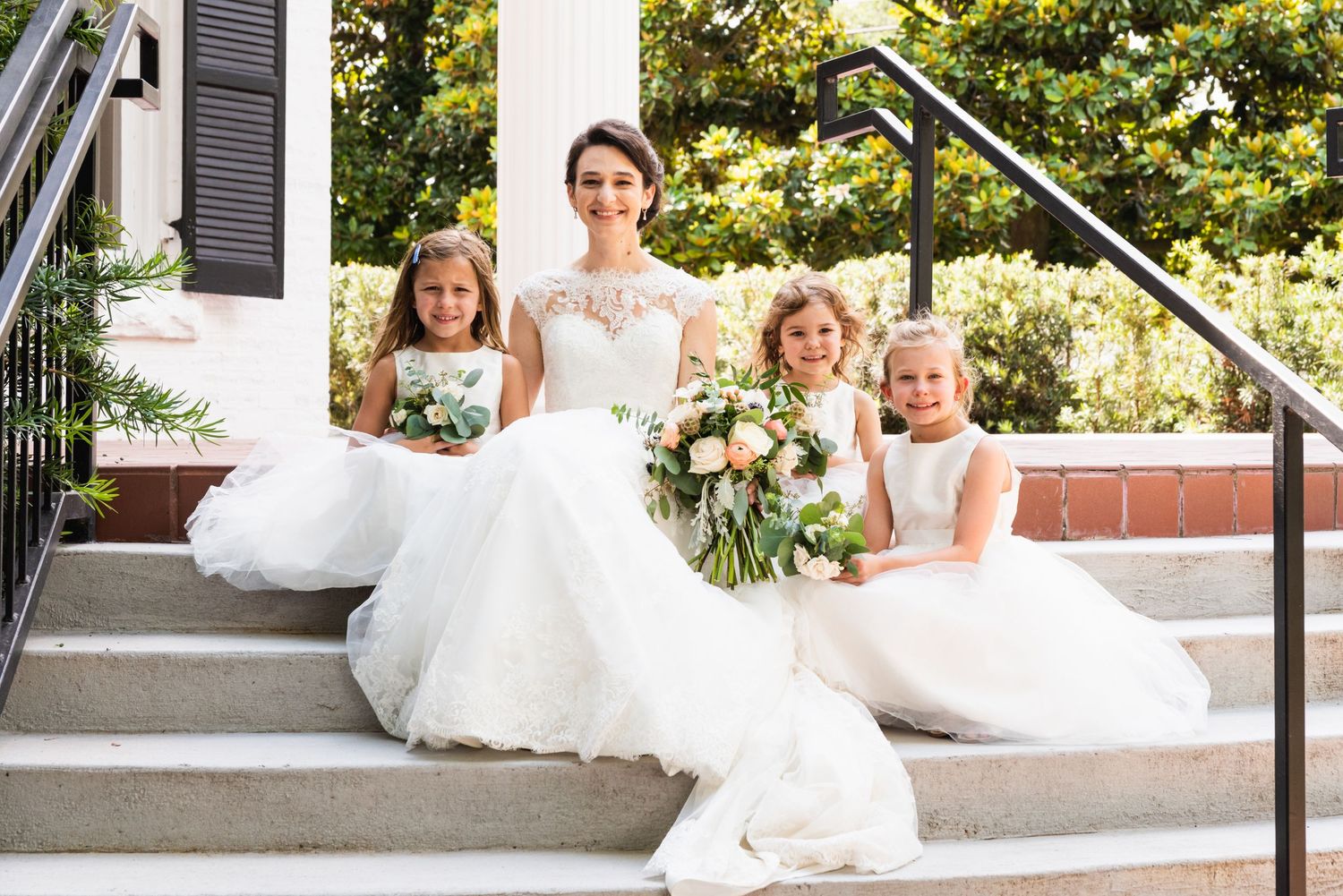 Bridal party in white dresses seated on outdoor mansion steps with floral bouquets.