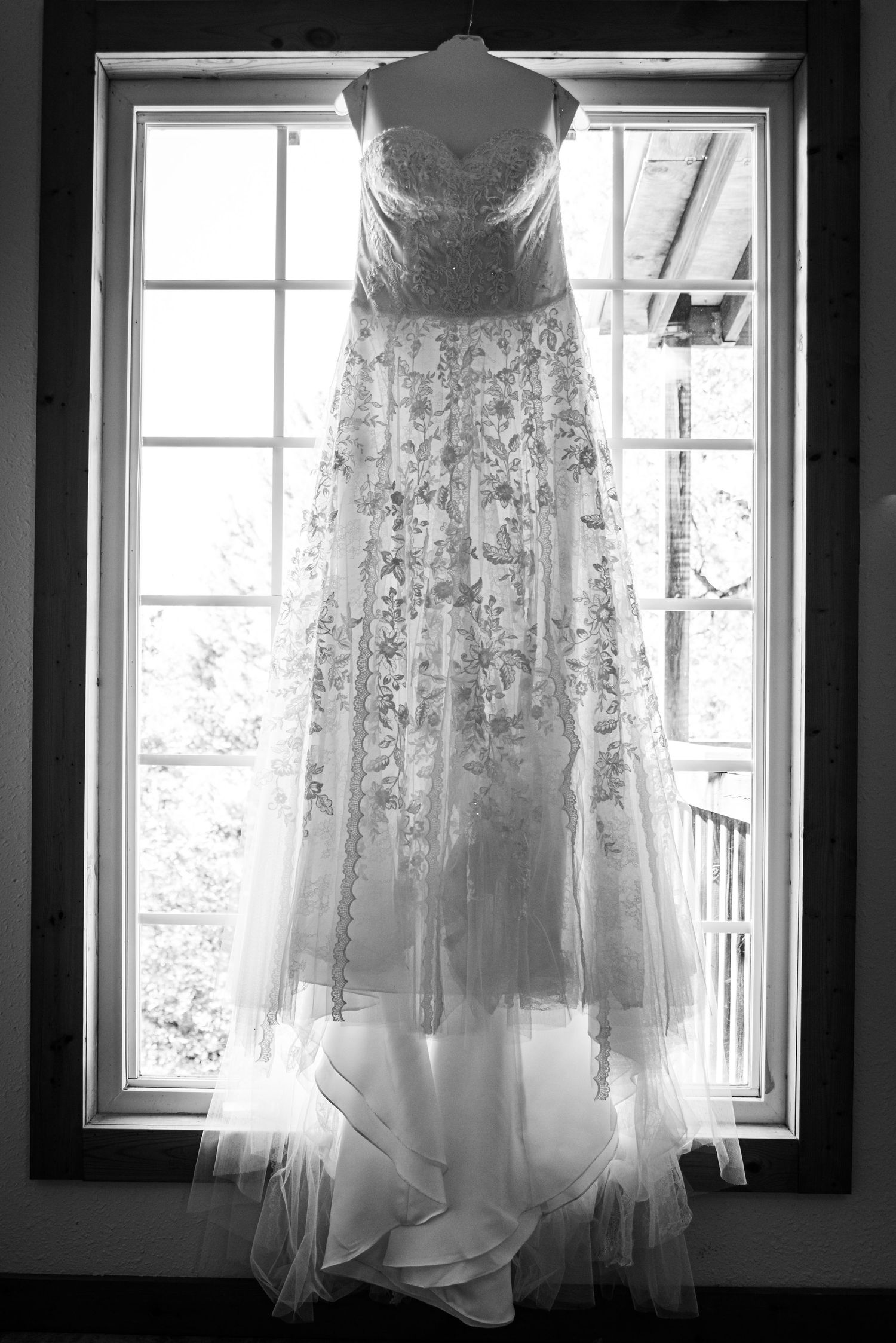 An elegant lace wedding dress hangs in front of a window in black and white.